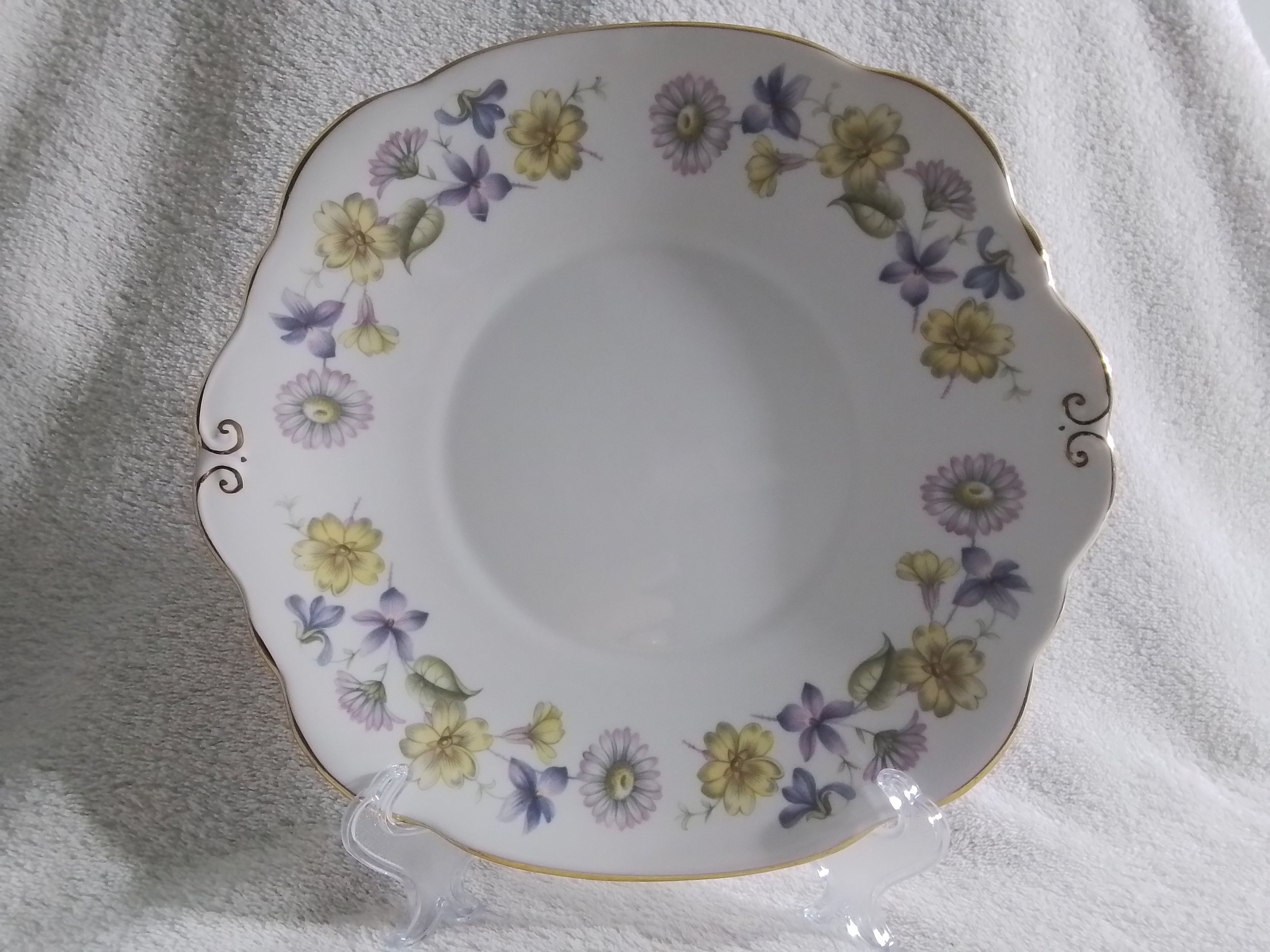 White porcelain plate with a scalloped edge and a floral pattern of yellow, purple, and lavender flowers, with a gold rim, displayed on a clear stand on a light-colored fabric background.