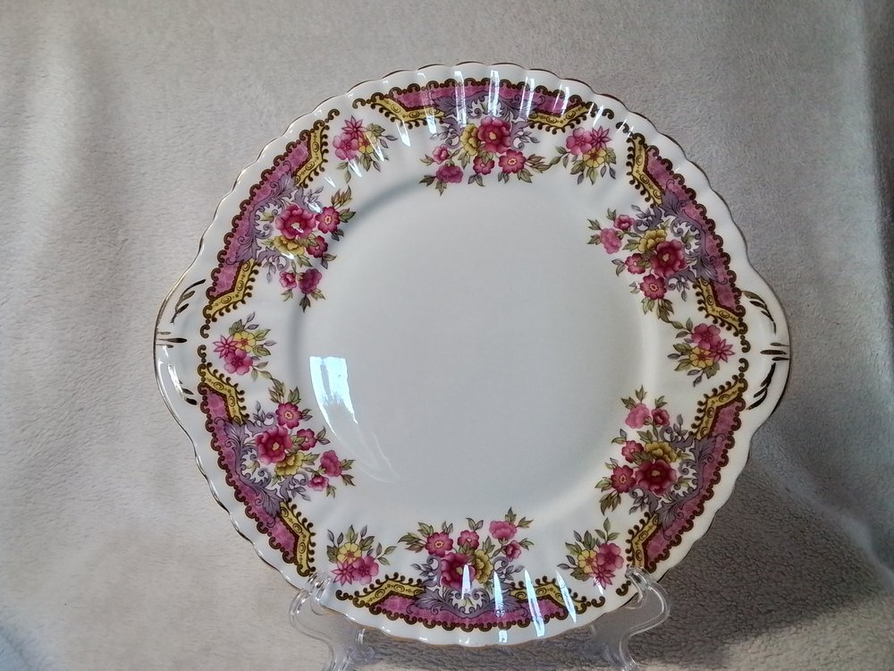 Decorative porcelain plate with pink, purple, and yellow floral pattern along the scalloped edge, displayed on a clear stand against a gray fabric background.