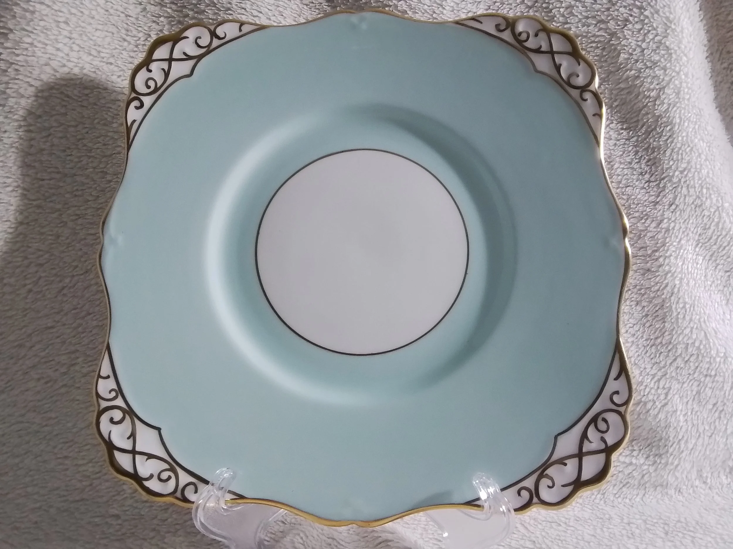 A decorative porcelain plate with a light blue center, gold and black ornate border, displayed on a clear stand, resting on a soft beige fabric background.