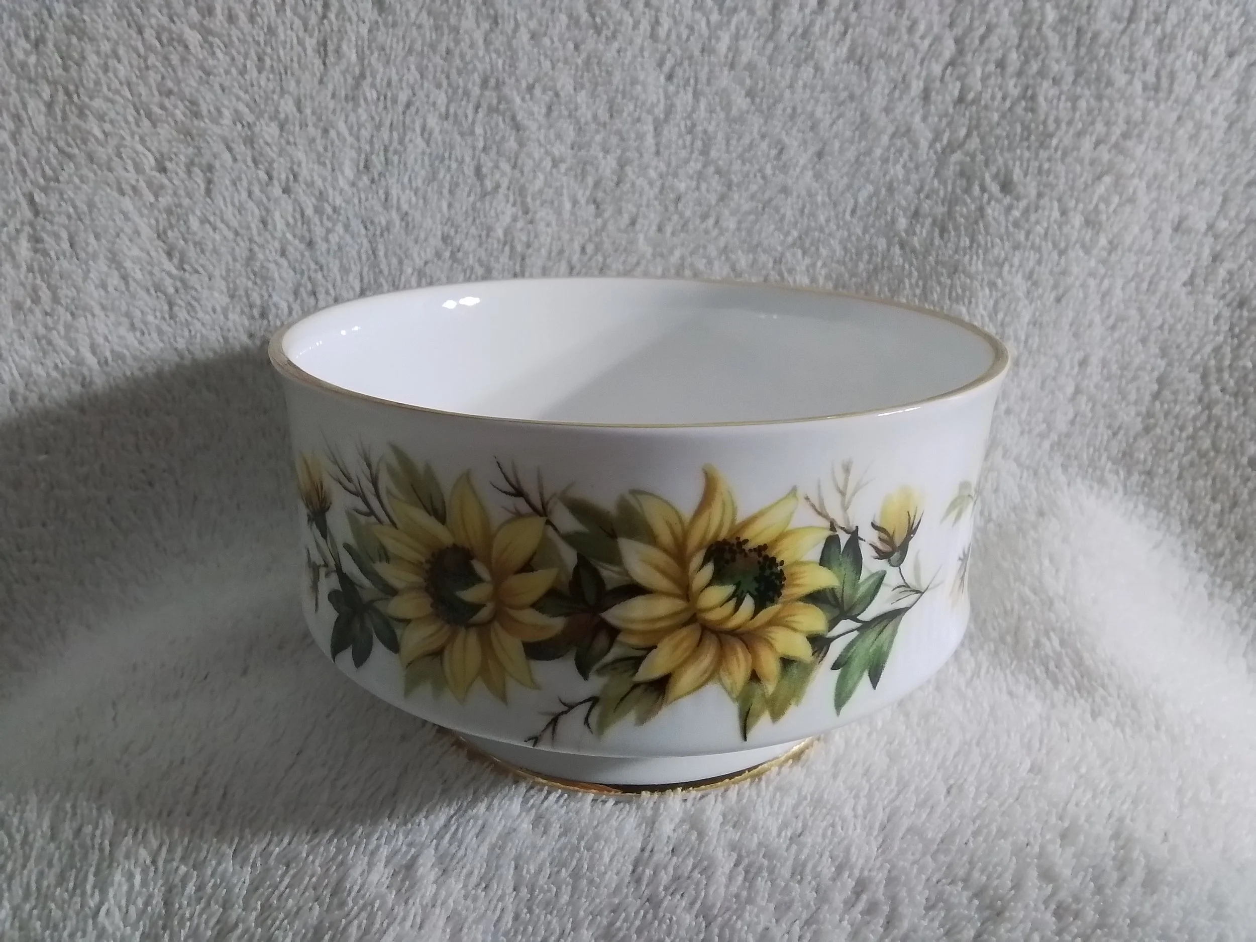 A white ceramic bowl with sunflower and leaf pattern, gold rim, on a soft, textured, light-colored surface.