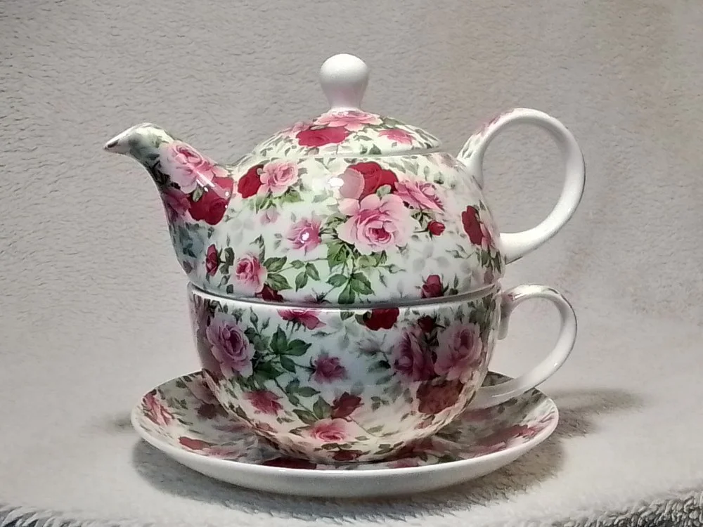 A floral-patterned teapot with a matching cup and saucer, all decorated with pink and red roses, placed on a cloth surface.