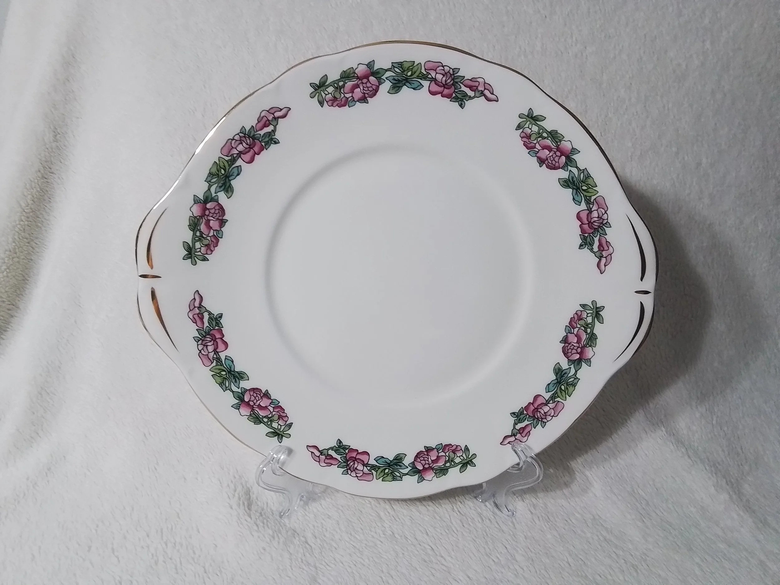 White ceramic plate with pink and green floral border, displayed on a clear stand on a white textured surface.