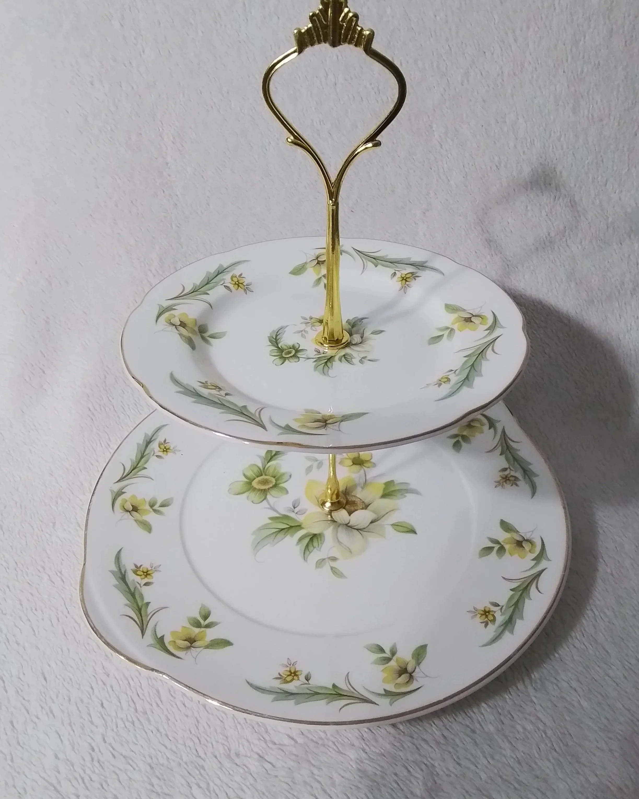 Two elegant porcelain dessert plates with yellow floral and green leaf patterns, stacked with a gold metal handle in the center for serving or display, placed on a white textured surface.