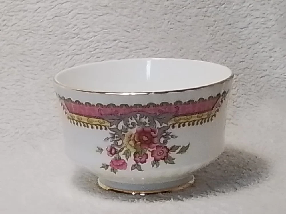 A decorative porcelain bowl with pink, grey, and gold floral patterns and a gold rim, placed on a textured white surface against a textured white background.