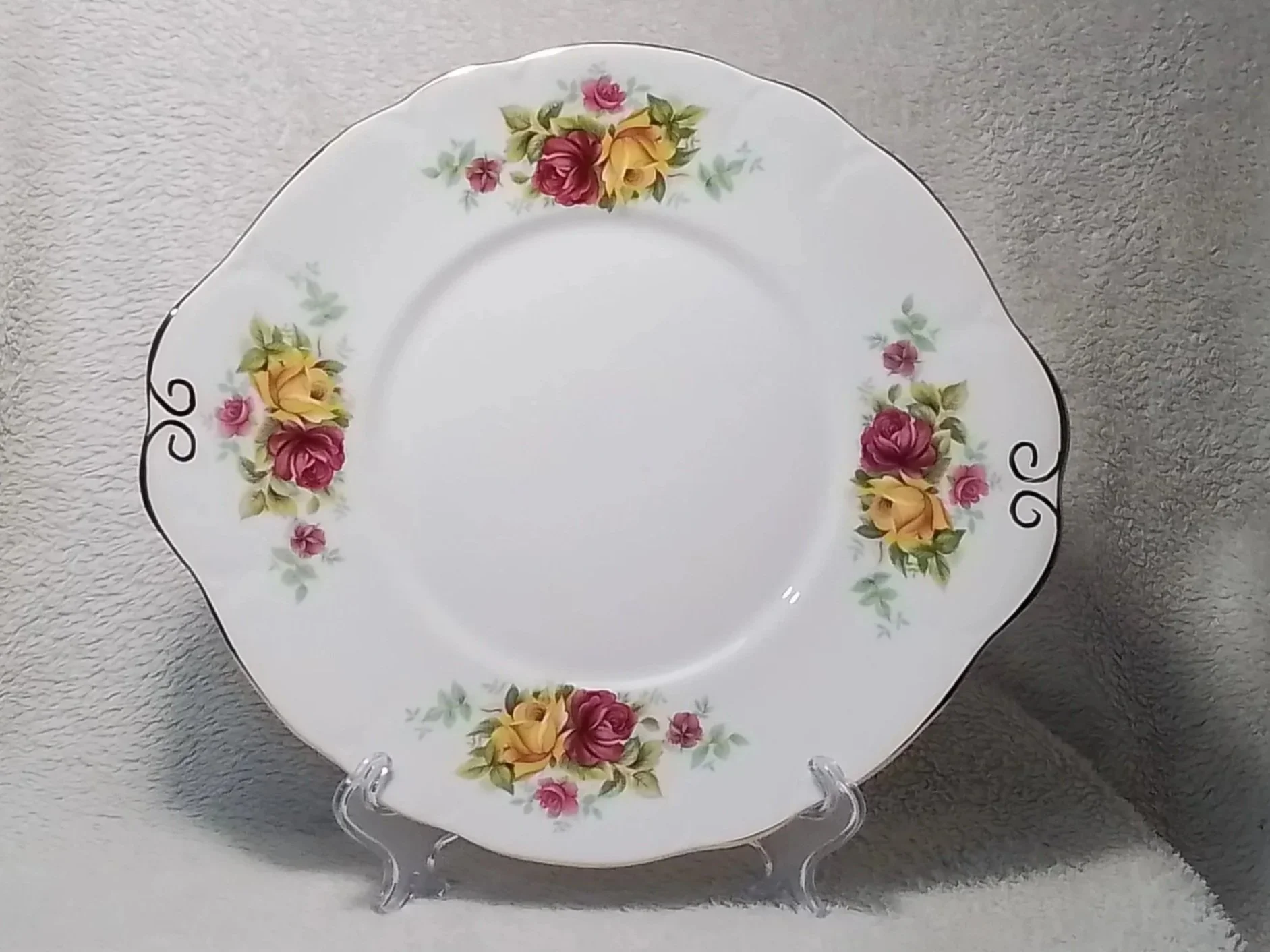 A decorative porcelain plate with a floral pattern of yellow and pink roses and green leaves, displayed on a clear stand against a gray textured background.