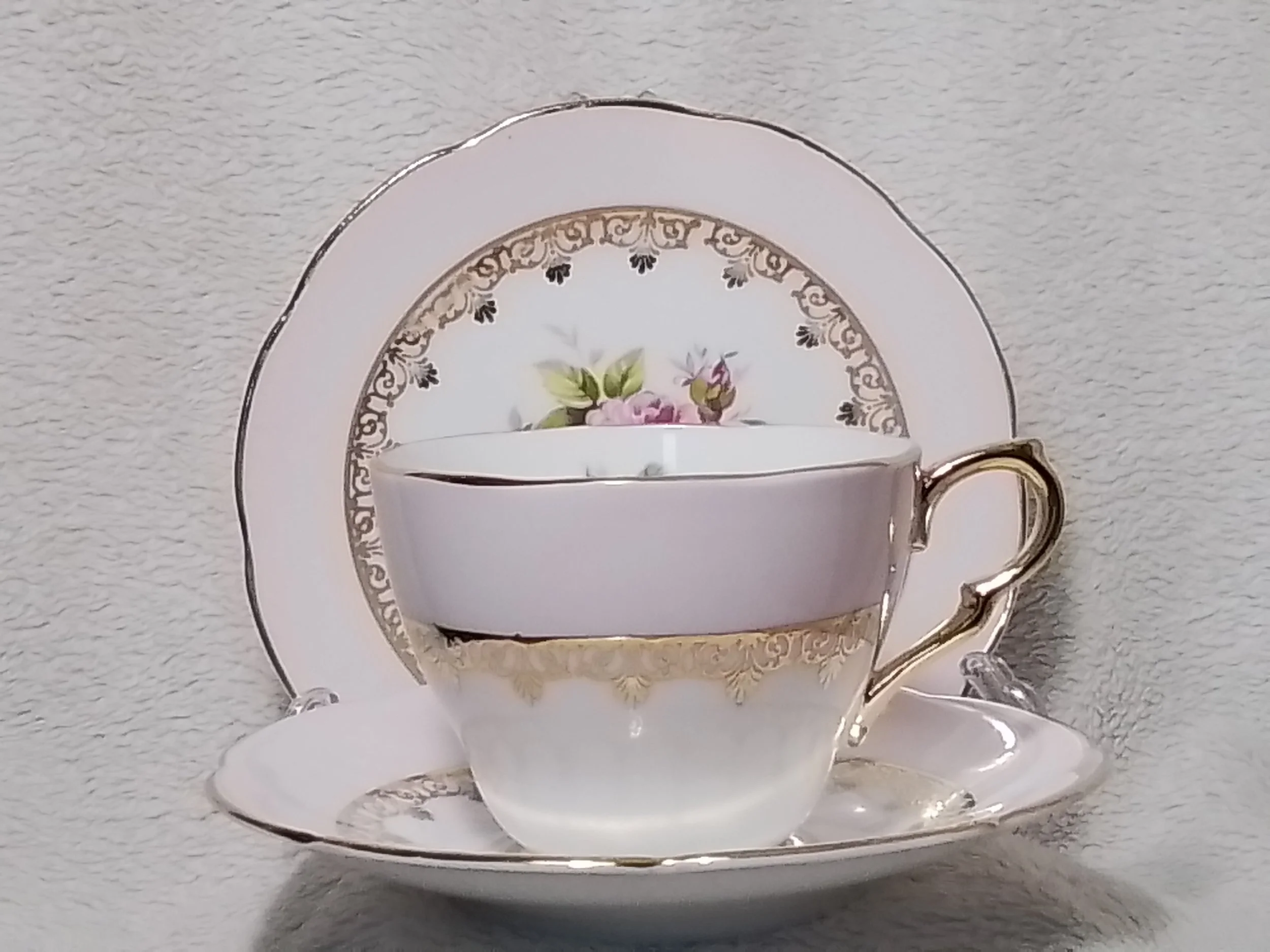 A delicate china teacup with gold trim and floral design, placed on a matching saucer with a patterned border, set against a textured gray background.