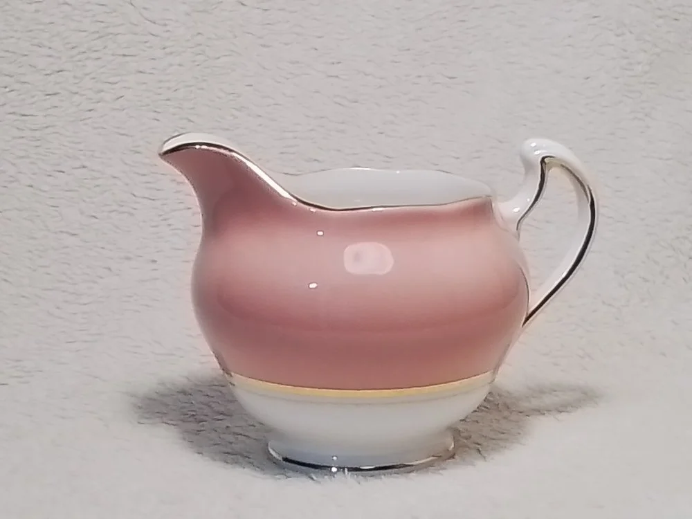 A pink and white porcelain creamer with a gold trim handle, placed against a textured white background.