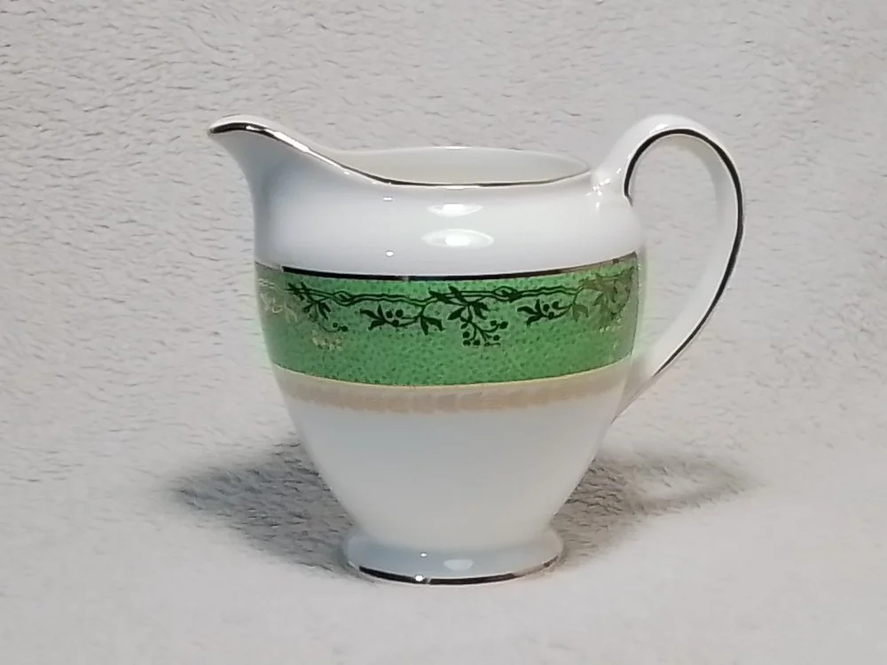 A white ceramic pitcher with green and black decorative trim, sitting on a light-colored surface.
