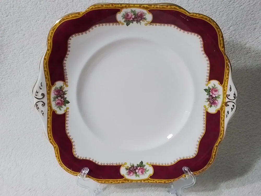Decorative porcelain plate with a floral pattern and gold trim, placed on a clear stand against a light-colored wall.