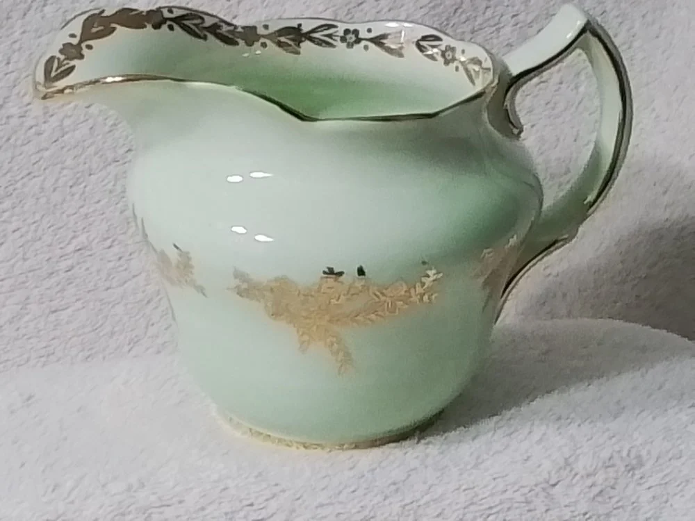 A vintage glass creamer with gold floral detailing around the rim, placed on a light-colored fabric surface.