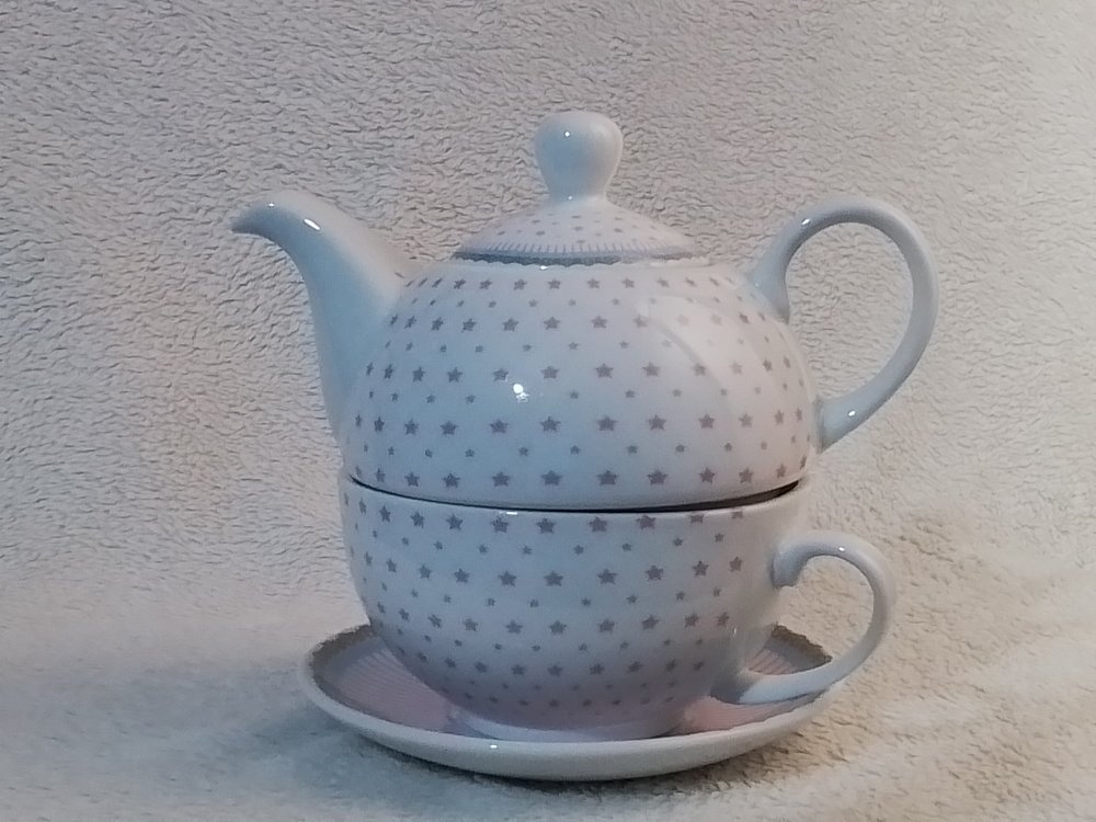 A white ceramic teapot with a lid and a matcha bowl with a handle, both decorated with small gray stars, stacked on a matching plate, set against a textured, light background.