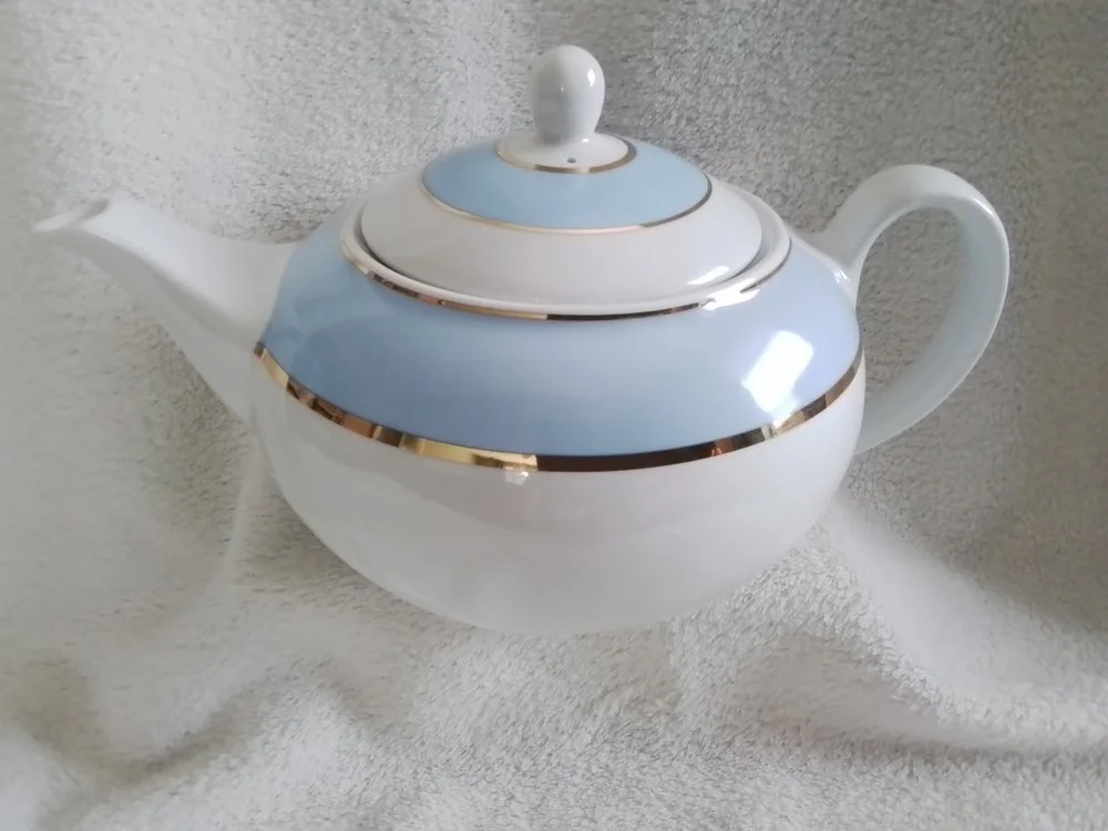 A porcelain teapot with a white, blue, and gold trim design on a soft, textured white surface.