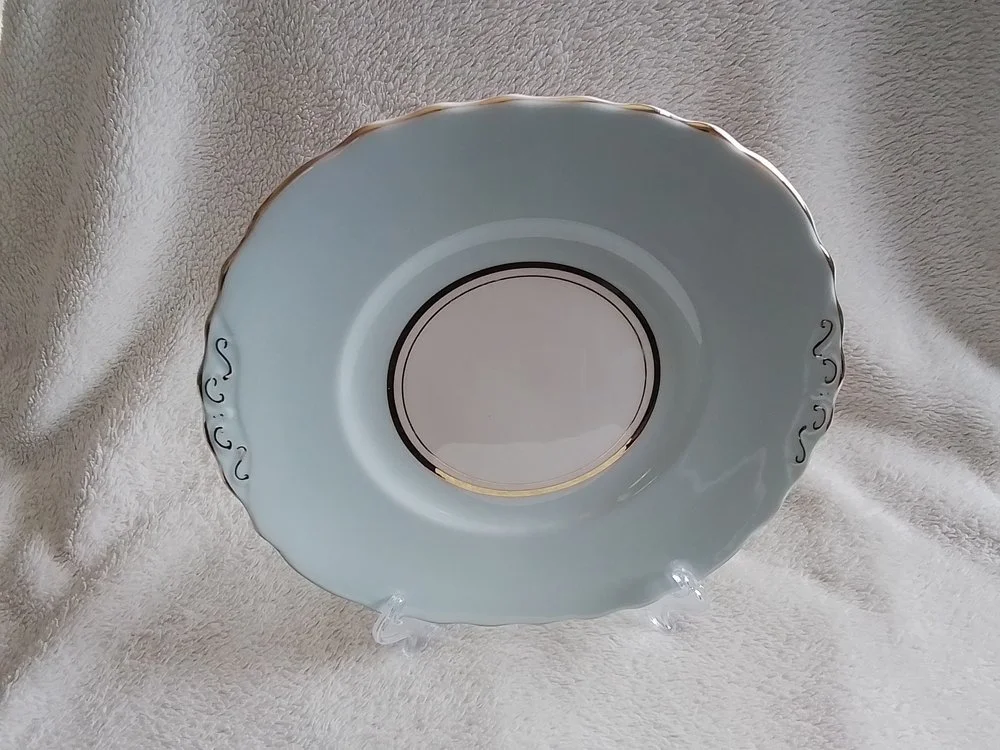 A white porcelain saucer with gold accents and decorative black scrollwork on the scalloped edges, displayed on a clear stand.