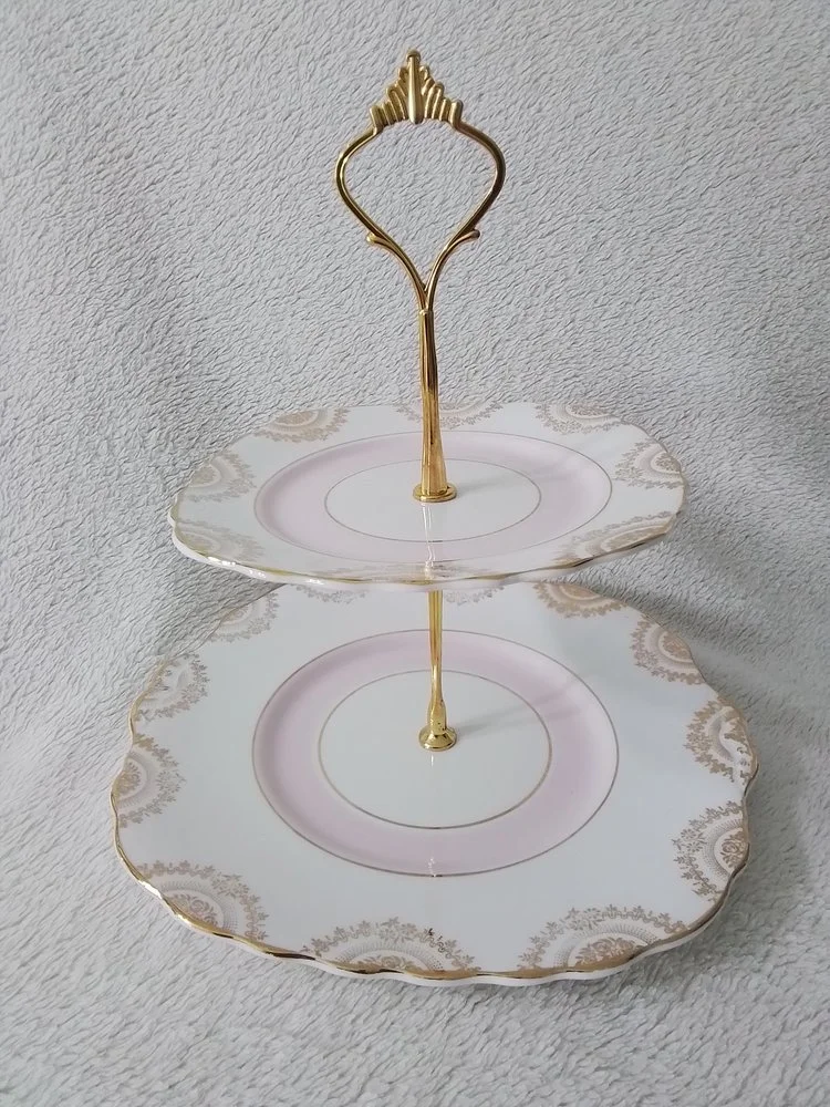Two-tiered decorative serving tray with white plates featuring gold and beige patterns, and a gold handle and support, placed on a gray textured surface.