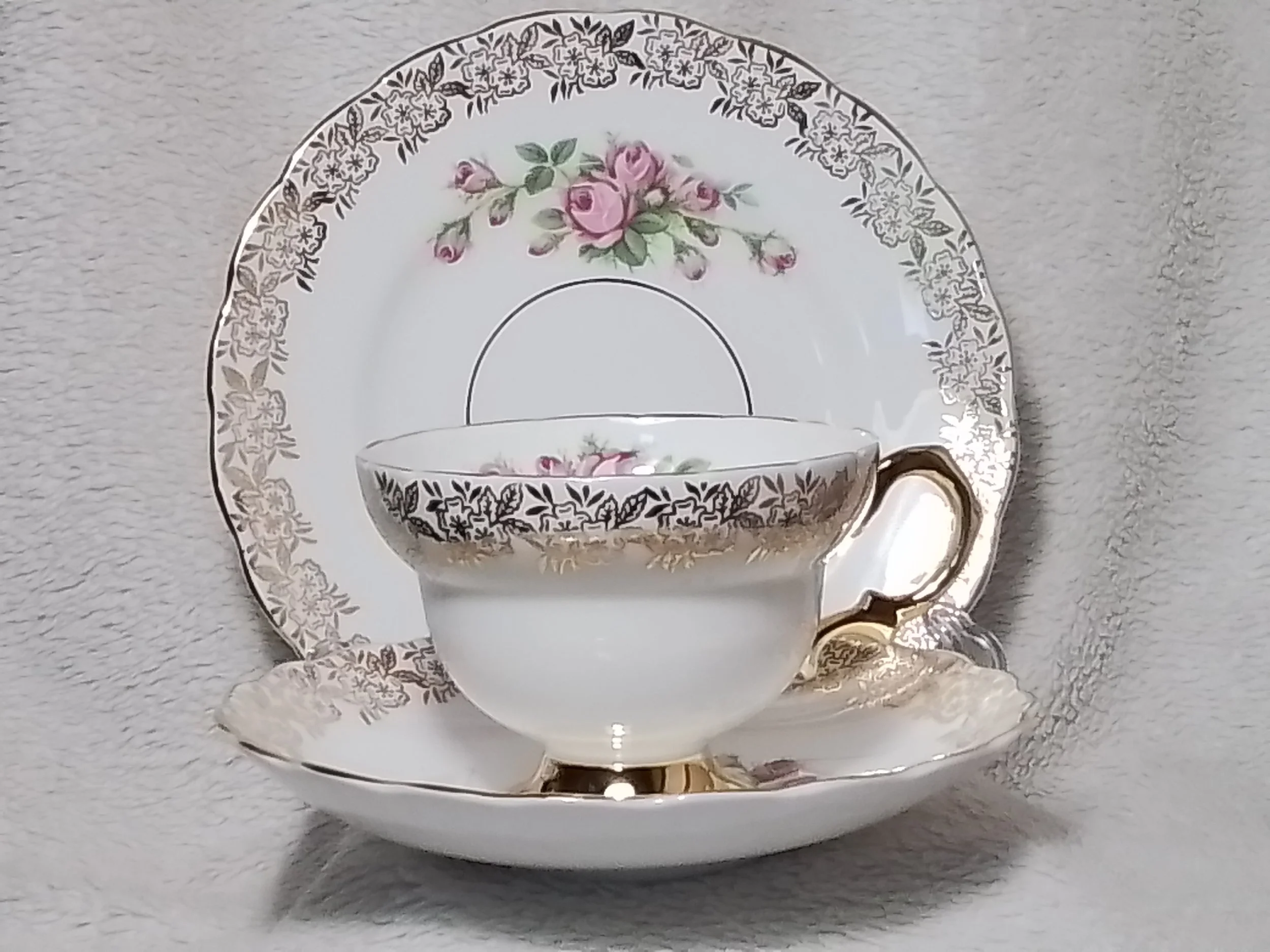 An ornate tea cup with a floral pattern and gold accents, placed on a matching saucer behind a large floral-patterned plate. The set features pink roses and intricate designs in gold and black.