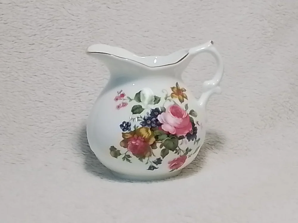 A small white ceramic pitcher with a floral pattern of pink roses, blue and yellow flowers, and green leaves, sitting on a light-colored textured surface.