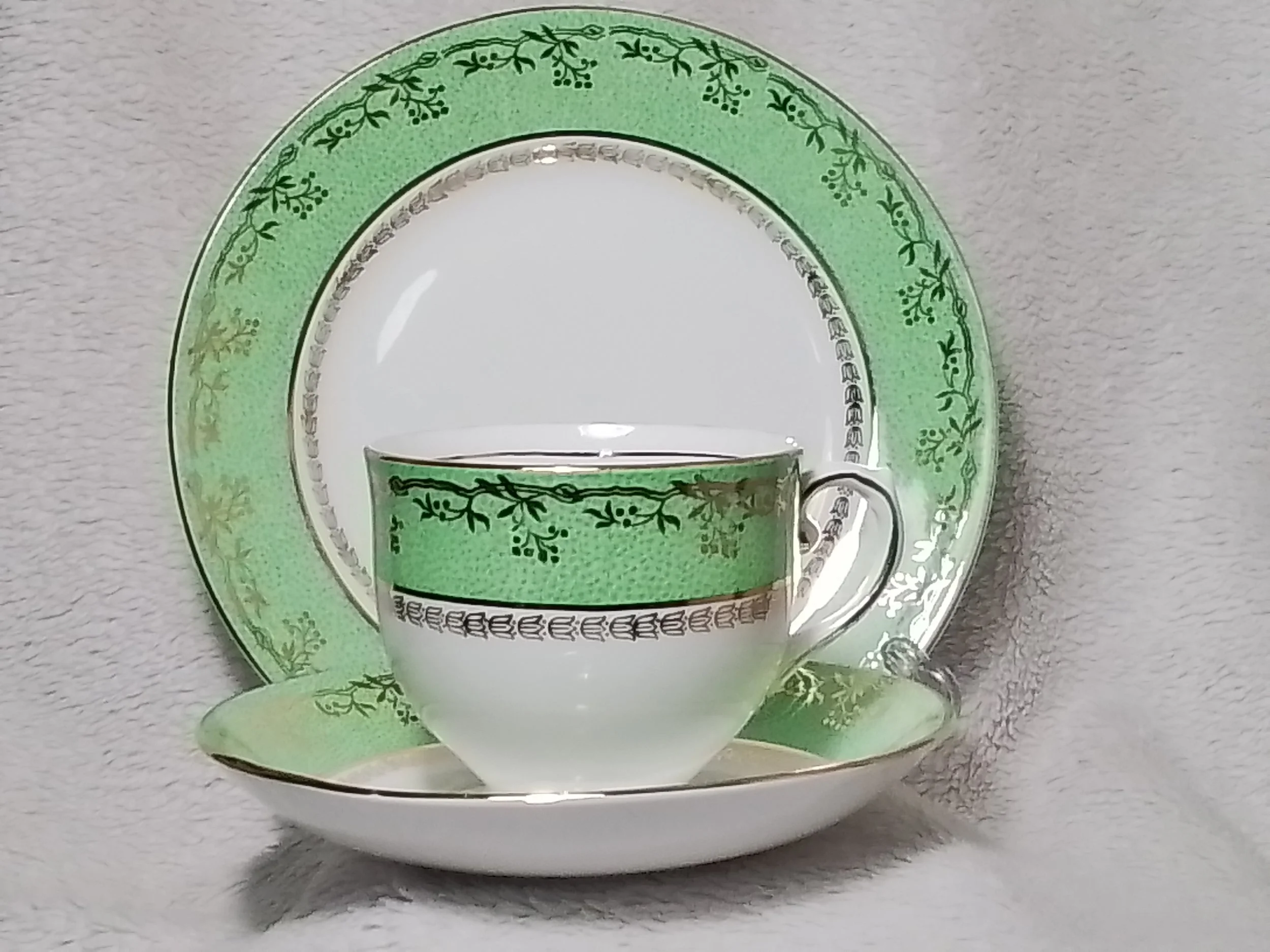A porcelain tea cup, saucer, and plate set with green and gold floral patterns on a light background.