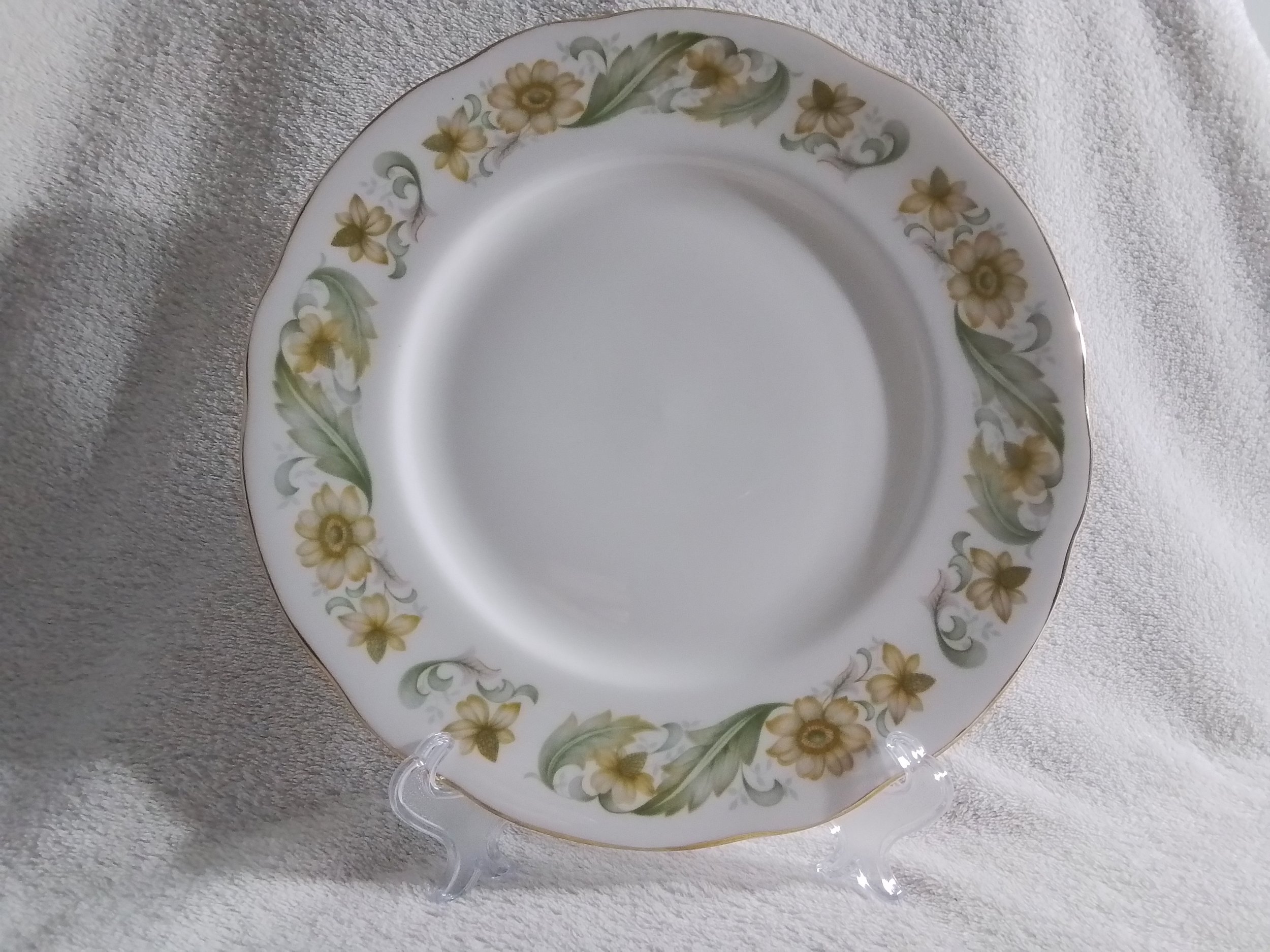 A porcelain plate with a floral pattern featuring yellow flowers and green leaves around the rim, displayed on a clear stand on a white textured surface.