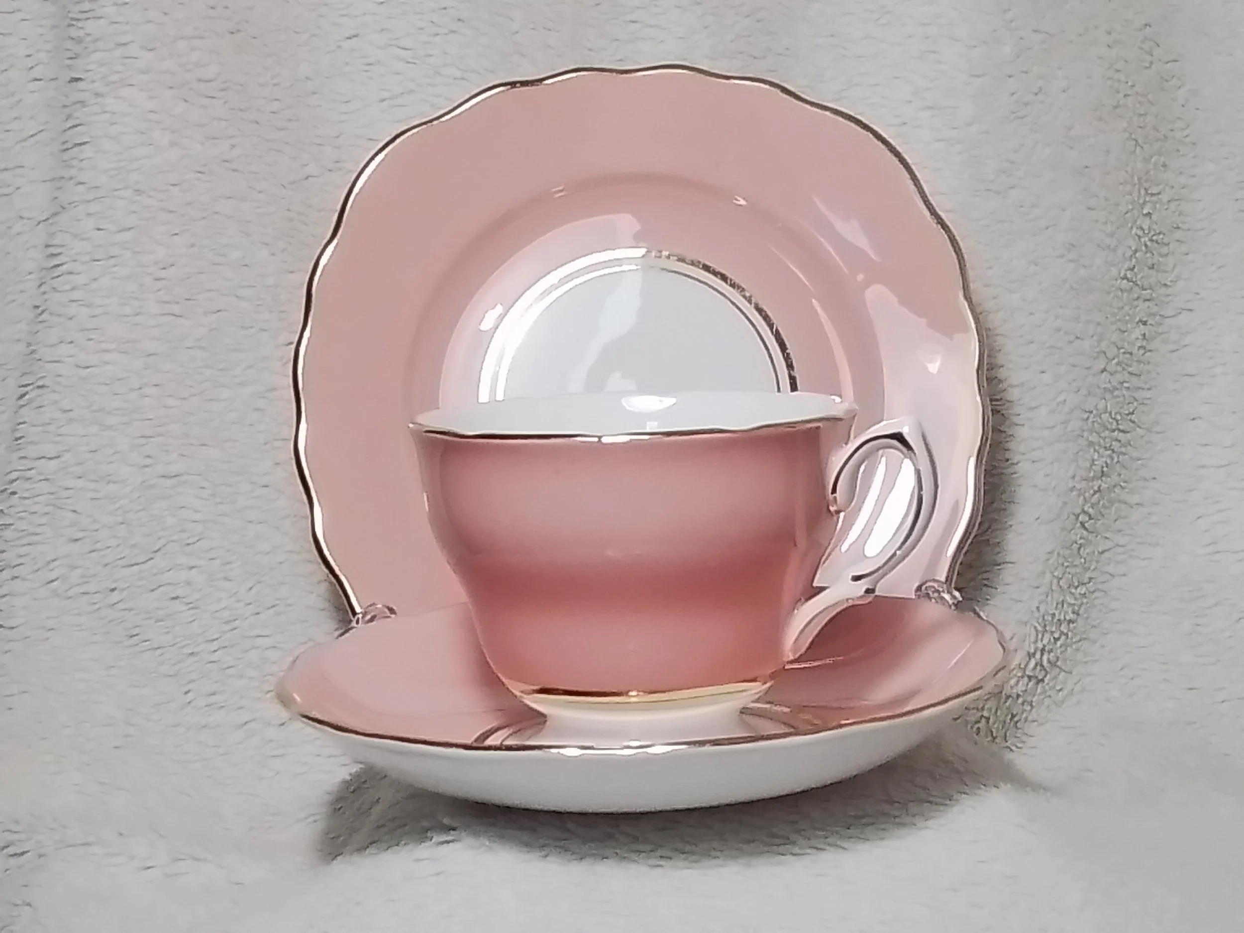 Pink teacup and saucer with gold accents, displayed on a matching pink plate with gold rim, on a light-colored textured surface.