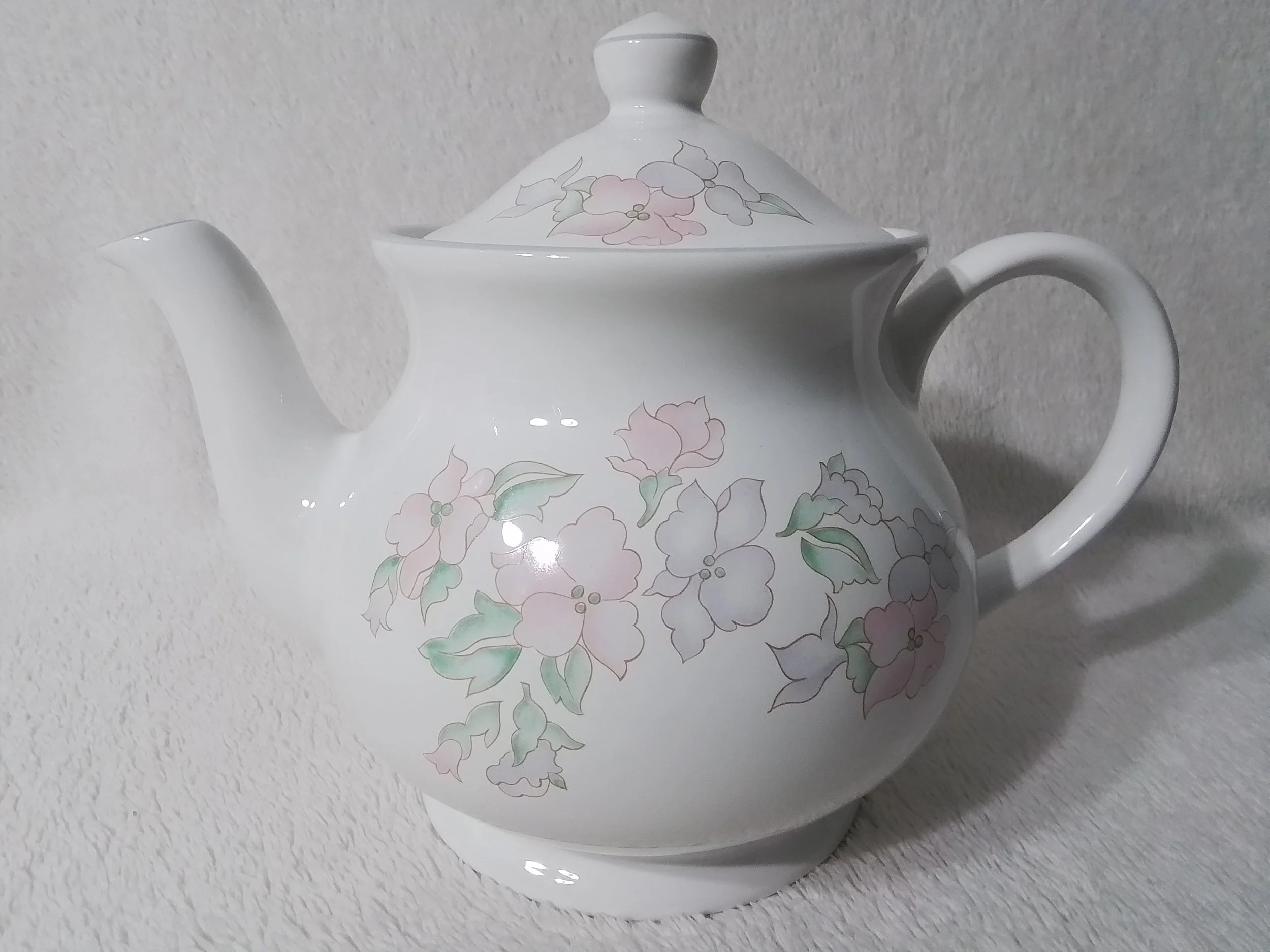 White ceramic teapot with floral pastel design and a lid, placed on a textured white surface.