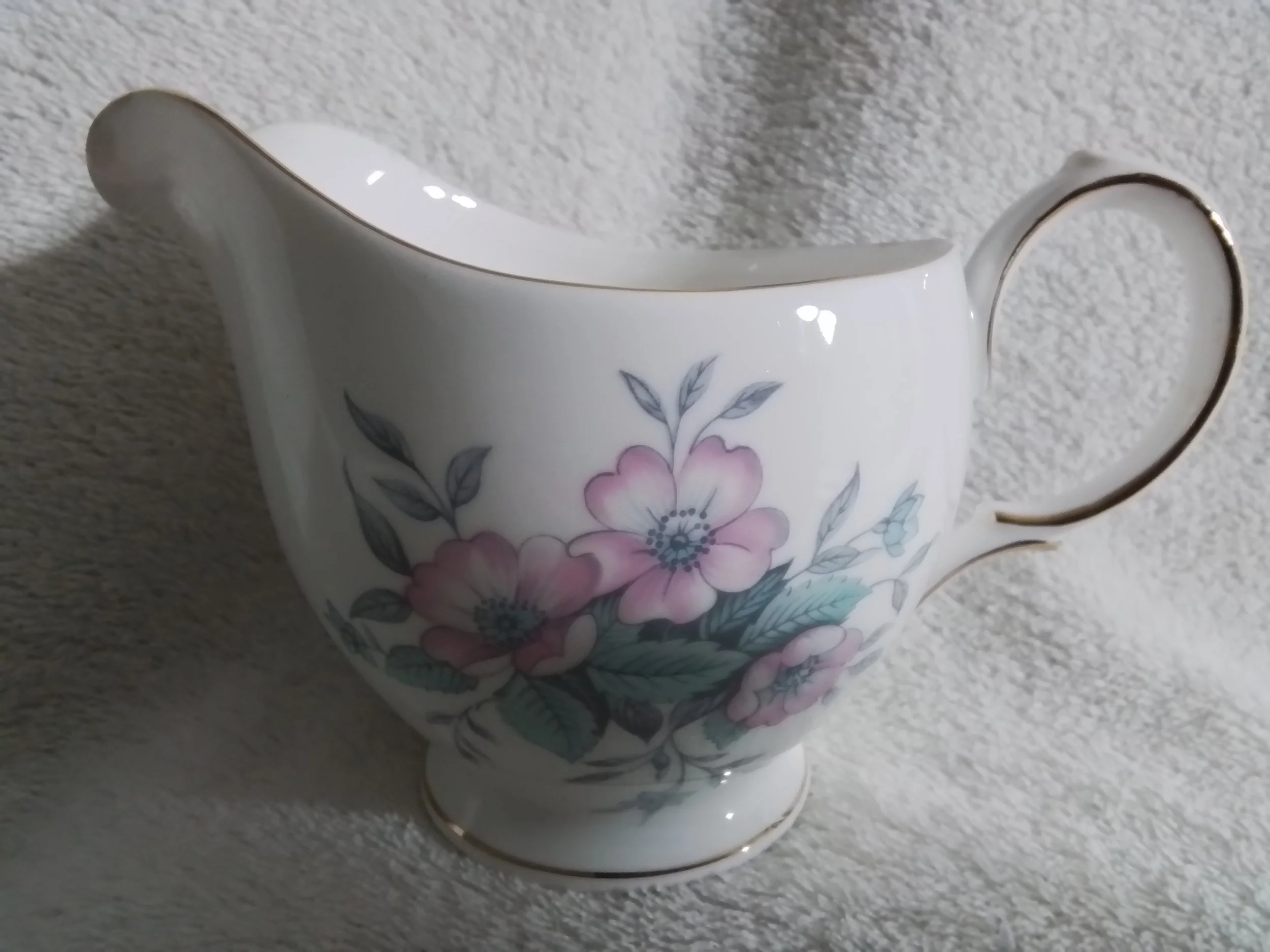 A white porcelain creamer with a floral design of pink flowers and gray leaves, featuring gold accents on the rim, handle, and foot.