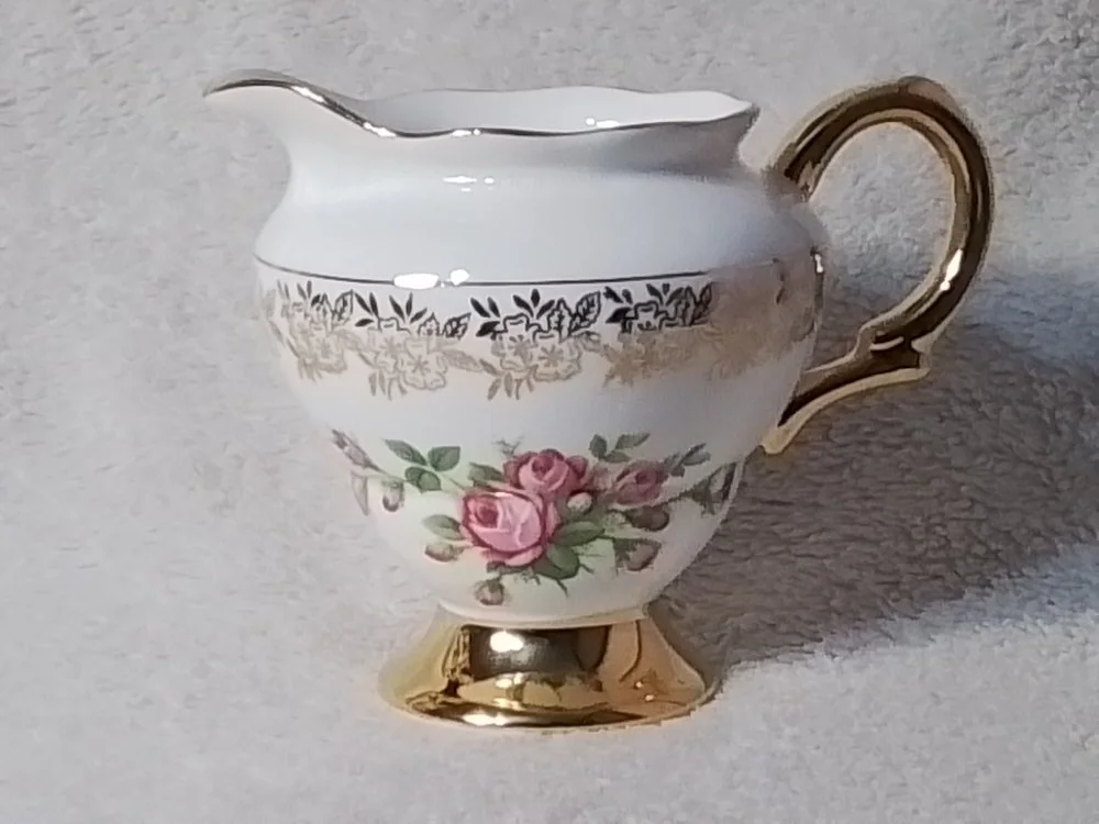 A decorative porcelain teapot with gold accents and a floral pattern of pink roses and gold detailing, placed on a light-colored surface.