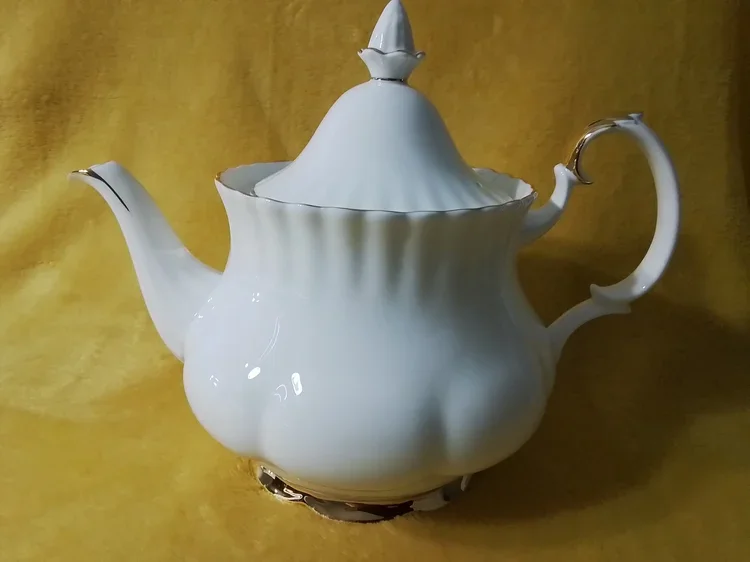 A white porcelain teapot with gold accents, ornate handle and spout, and a decorative lid, set against a yellow background.