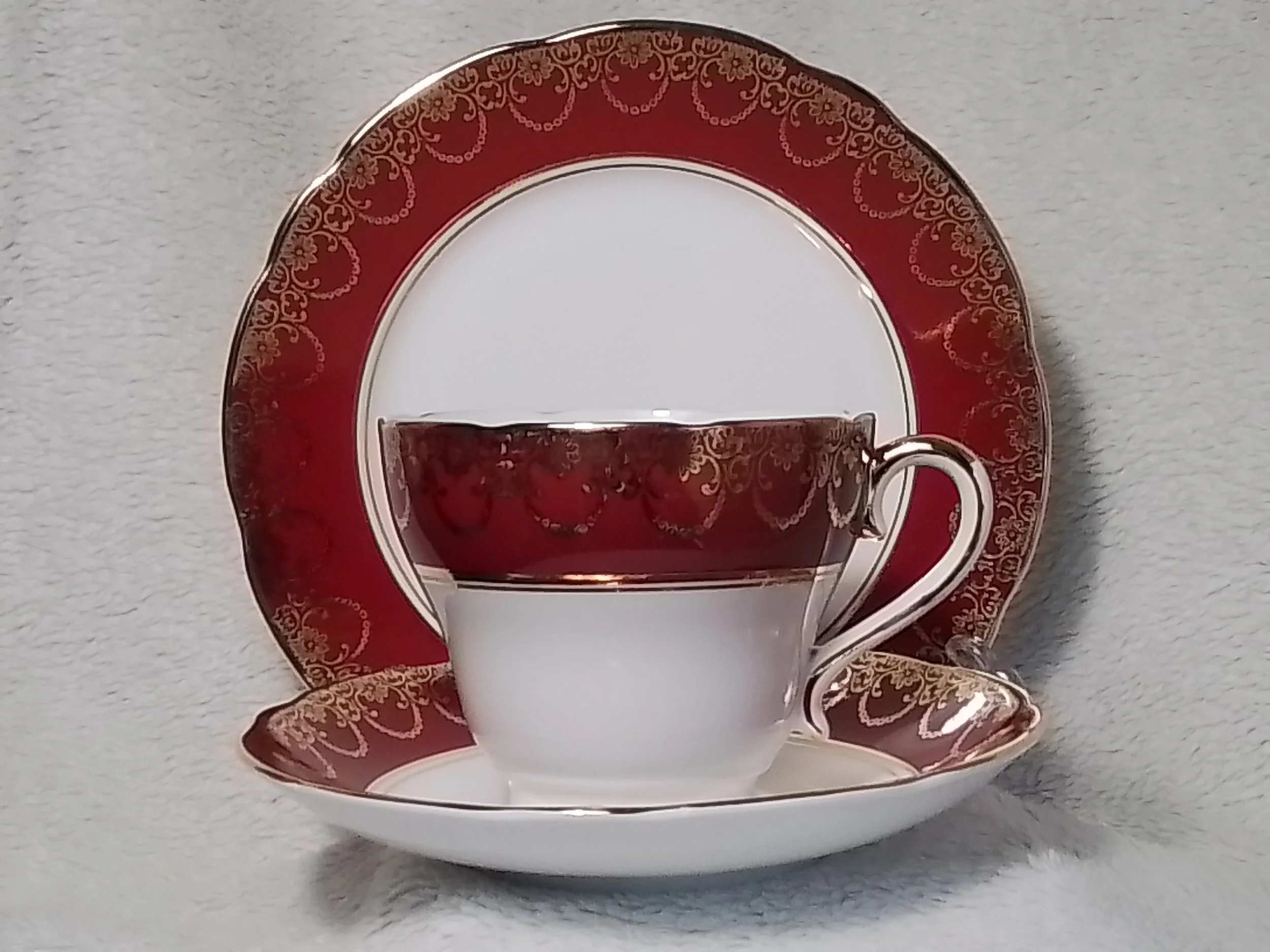A decorative tea cup with gold patterns, placed on a matching saucer with a matching plate behind it, all with a red and gold ornate design, set against a white background.