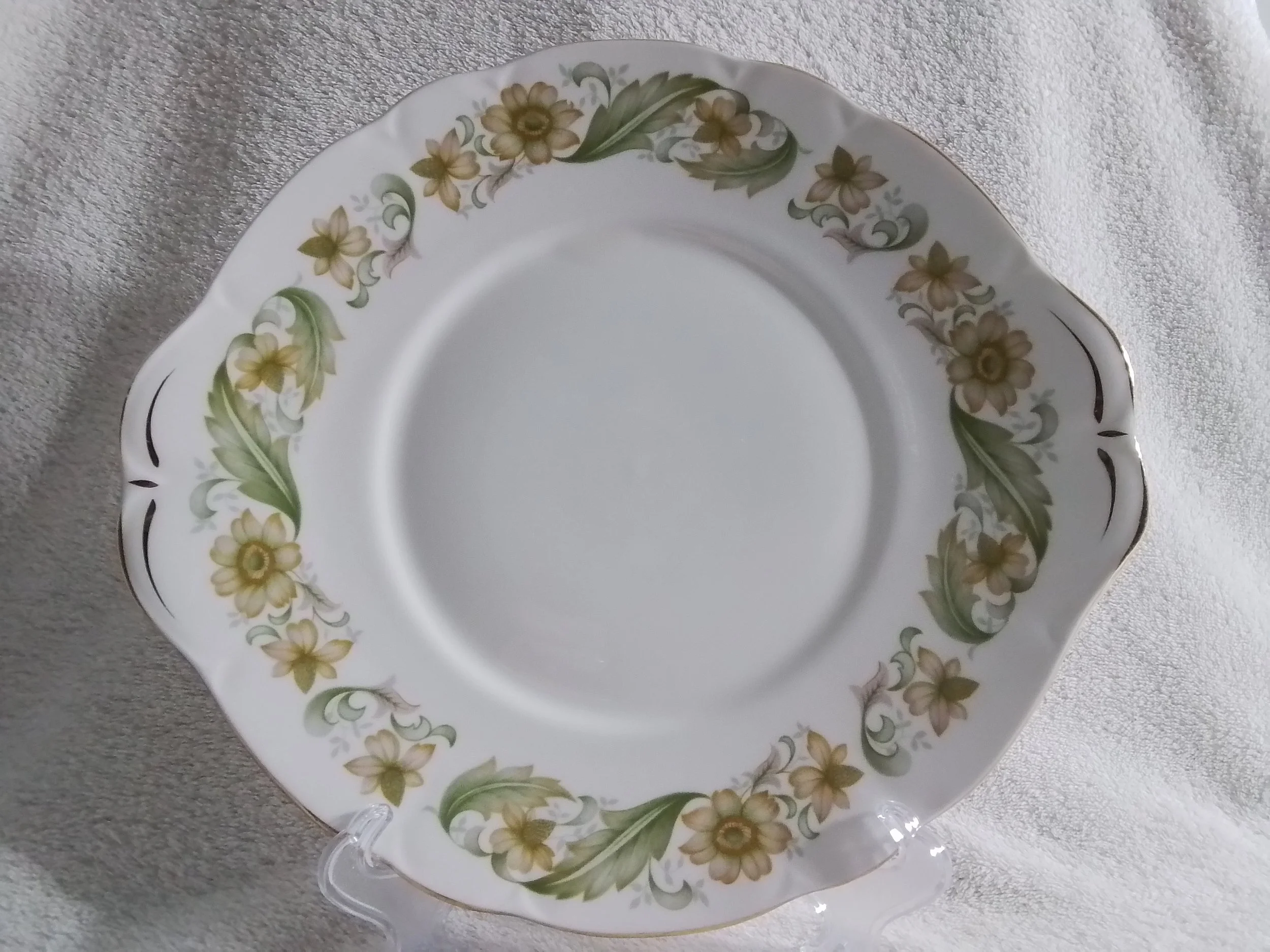 White decorative plate with a floral border featuring yellow and green flowers, gold accents, and a scalloped edge, placed on a light-colored fabric surface.