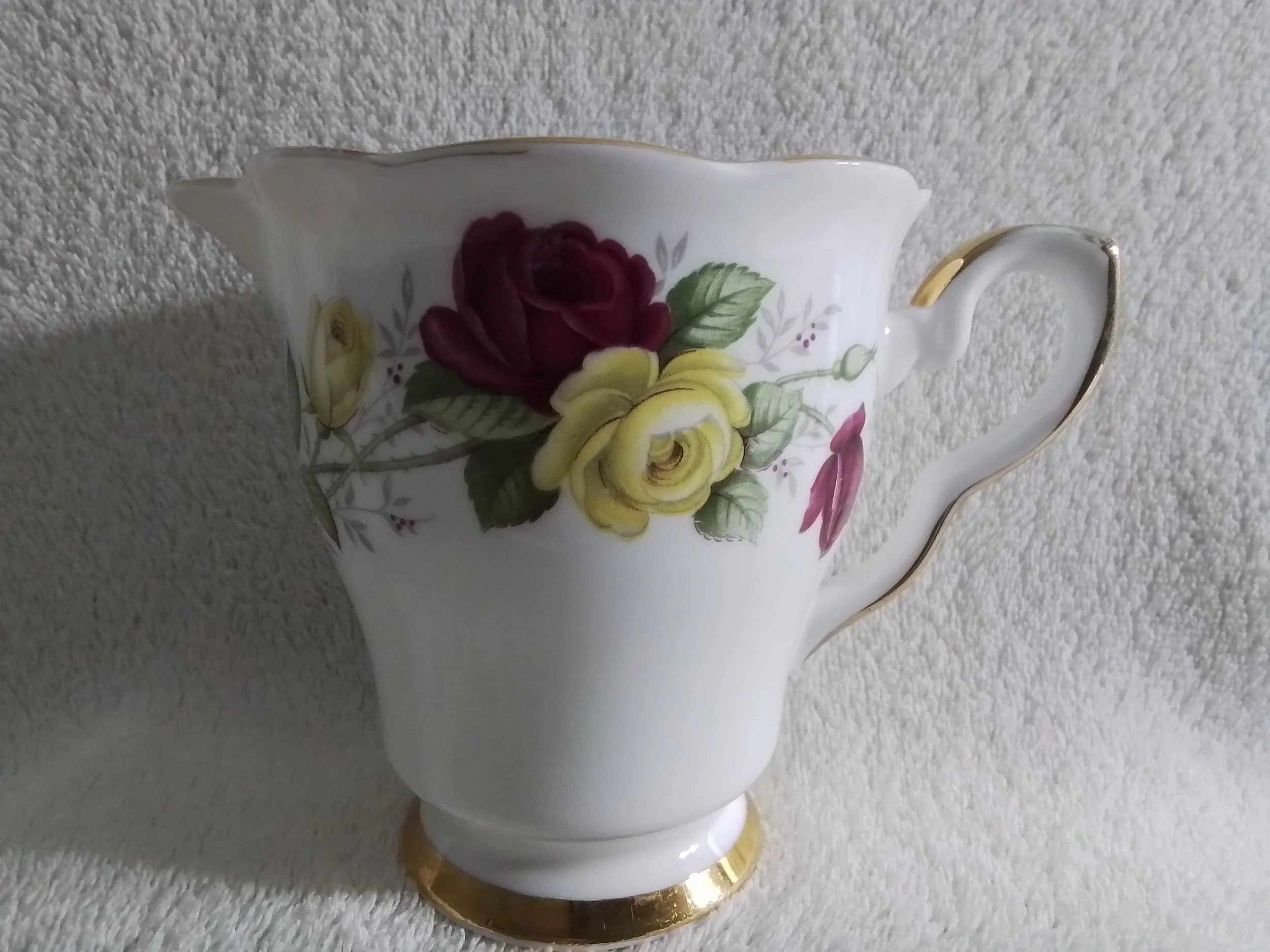 A porcelain teapot with gold trim and floral design featuring red, yellow, and pink roses against a textured background.