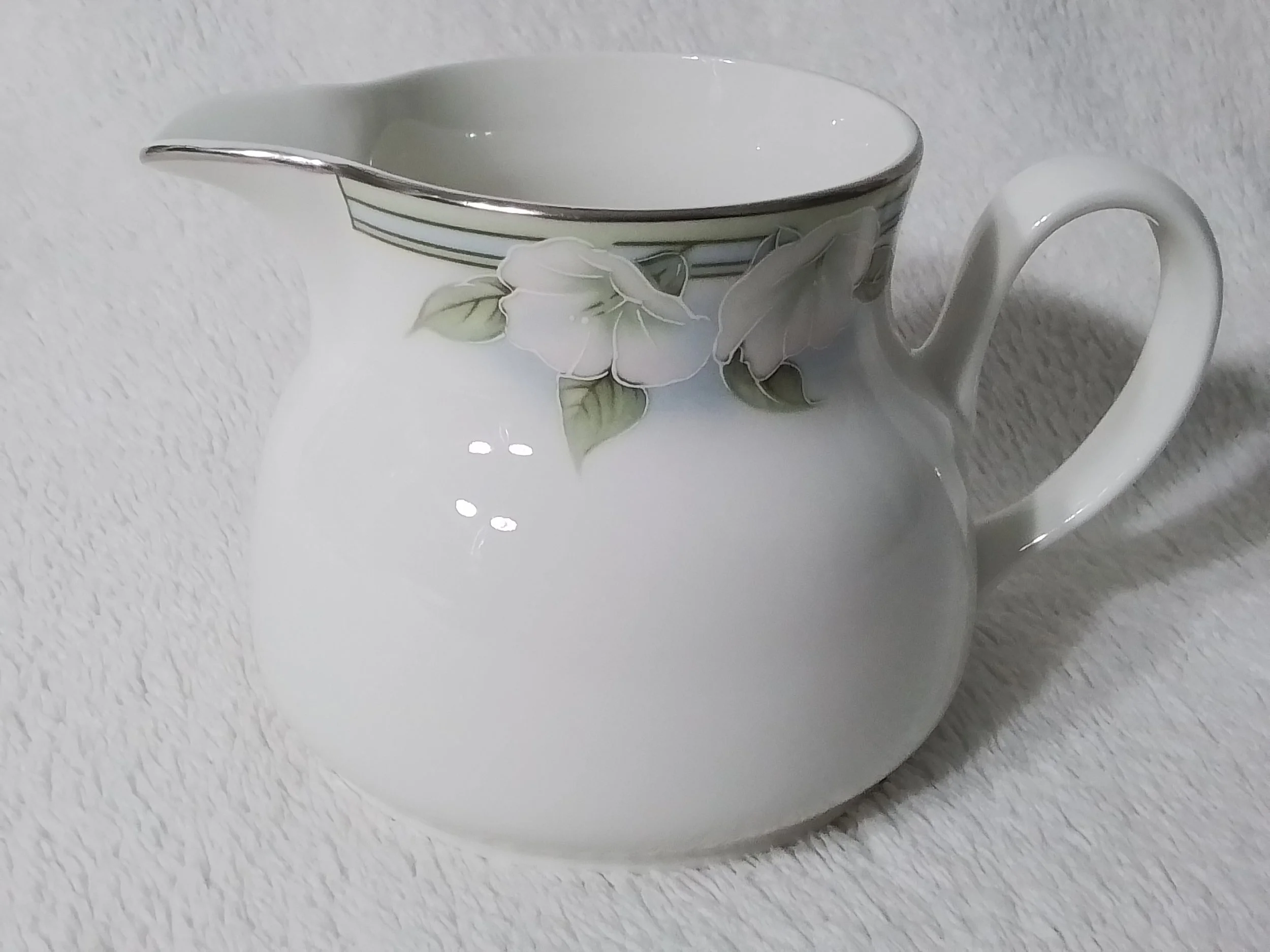 A white ceramic creamer with floral patterns and a silver rim, placed on a textured white surface.