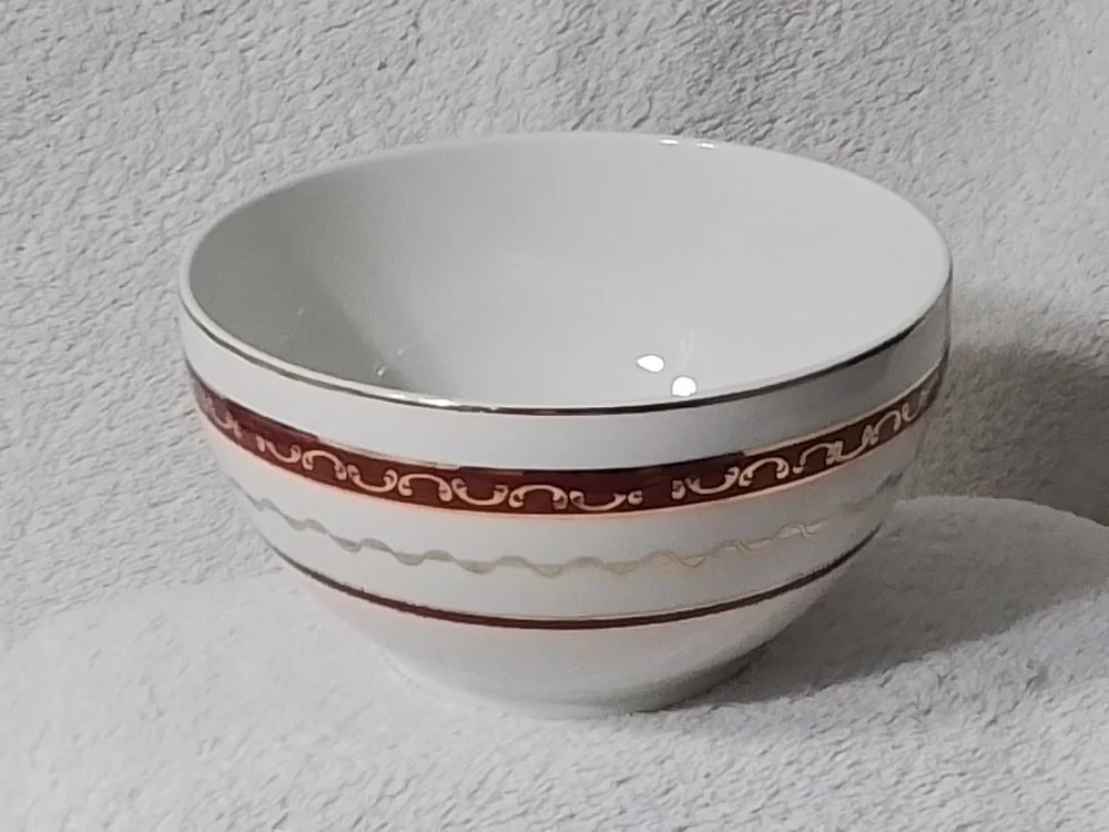 A white ceramic bowl with decorative red and gold bands around its exterior, placed on a light-colored textured surface.