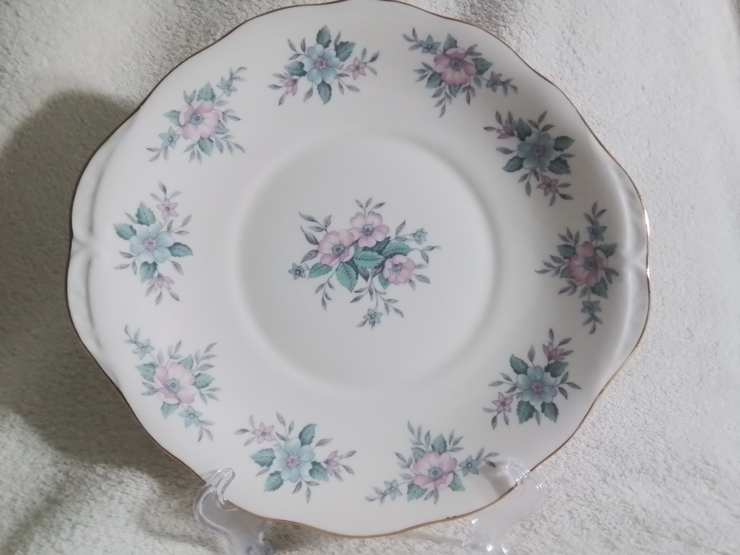 A white ceramic plate with scalloped edges and a floral pattern of pink, blue, and purple flowers with green leaves around the rim and center, placed on a clear plastic stand on a textured light-colored surface.