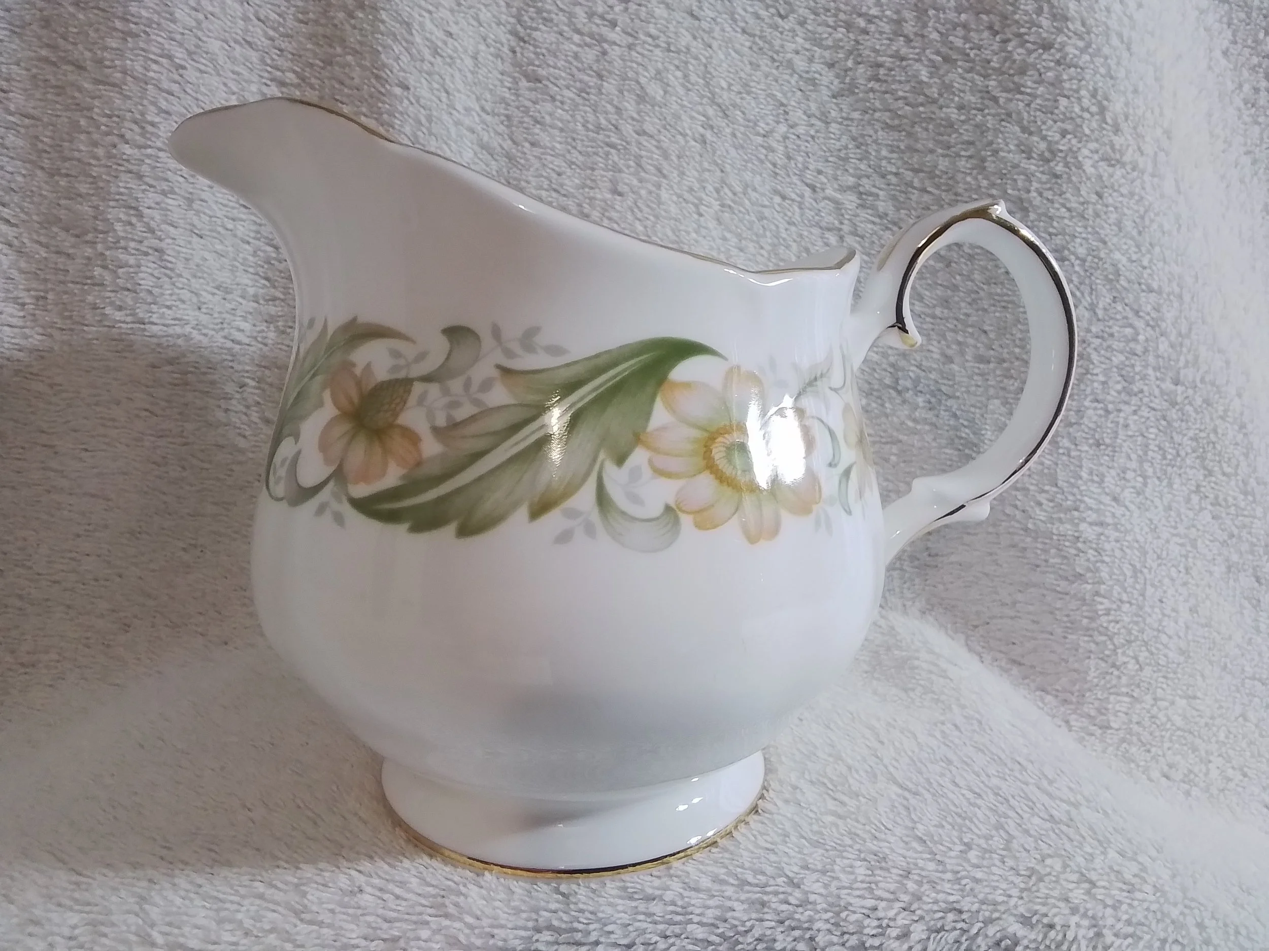 A white porcelain teapot with a floral pattern of yellow and orange flowers and green leaves, accented with gold trim, set on a beige textured surface.