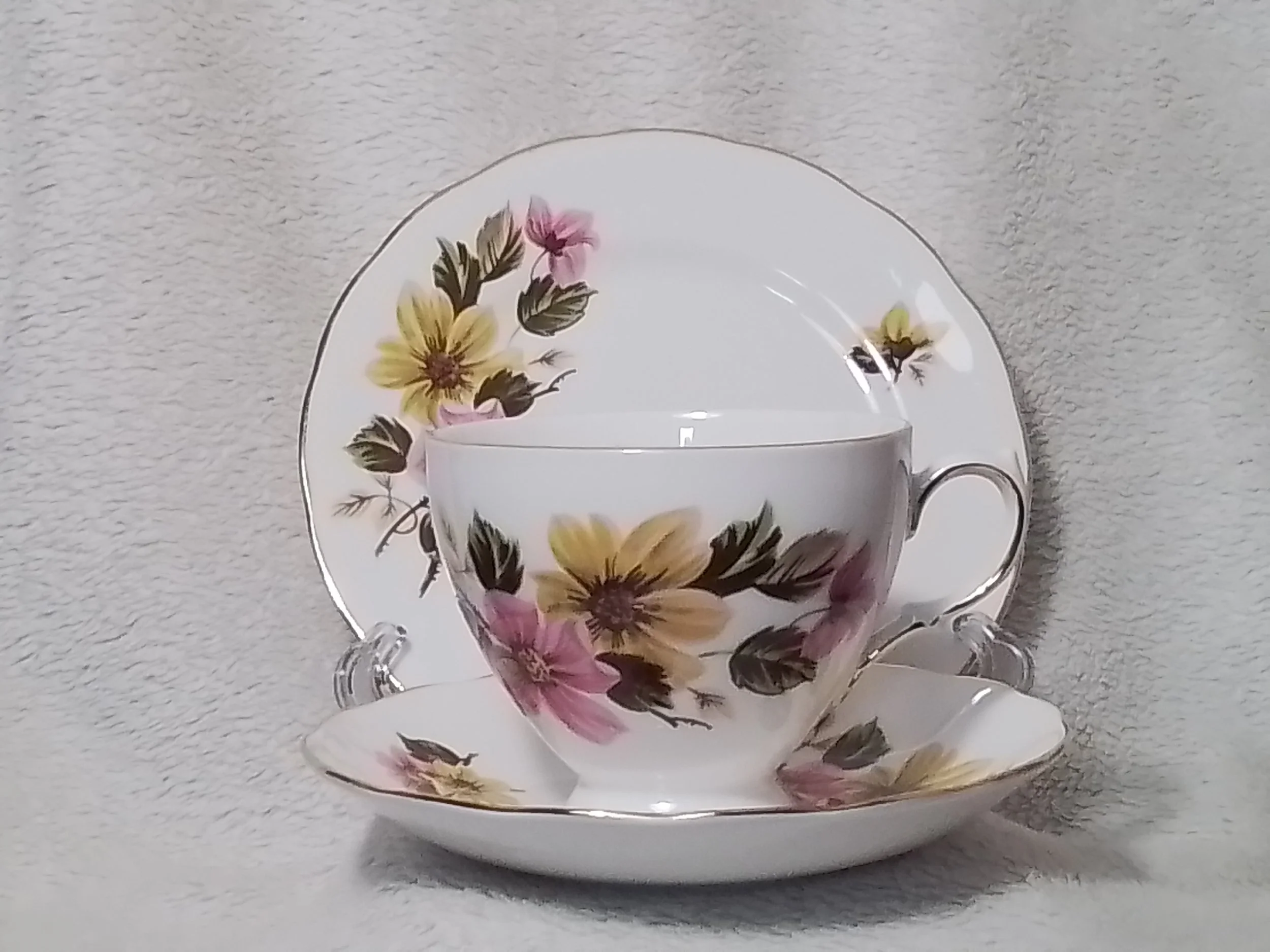 A floral patterned tea cup with matching saucer and plate, featuring yellow and pink flowers with green leaves, set against a plain background.