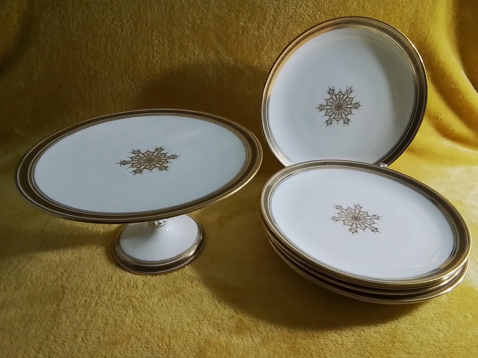 A set of fine china plates and a cake stand with gold and black decorative borders and intricate gold floral patterns in the center, displayed on a yellow fabric background.