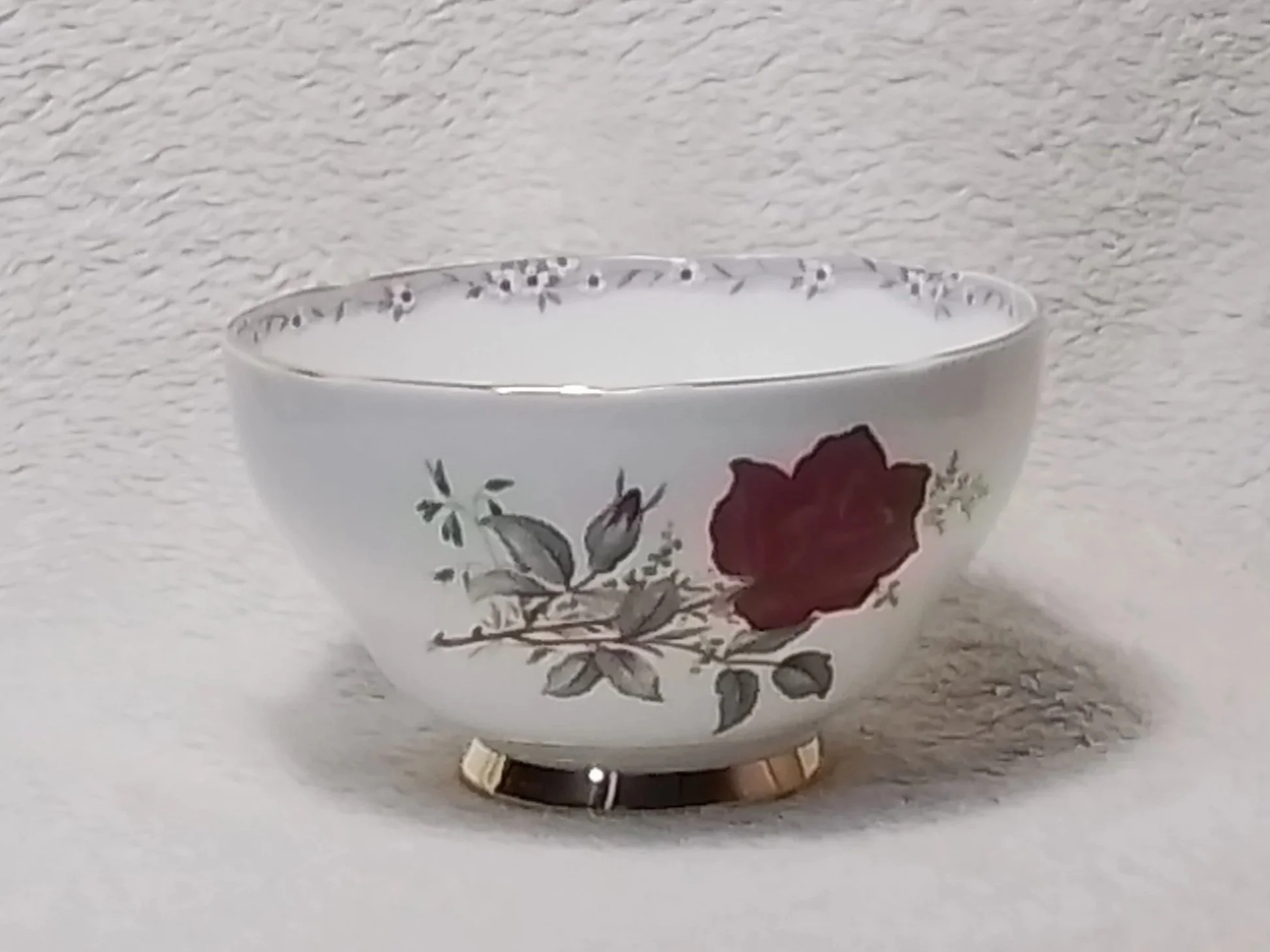 A white ceramic bowl with floral patterns, including a red rose and green leaves, placed on a white textured surface against a white textured background.