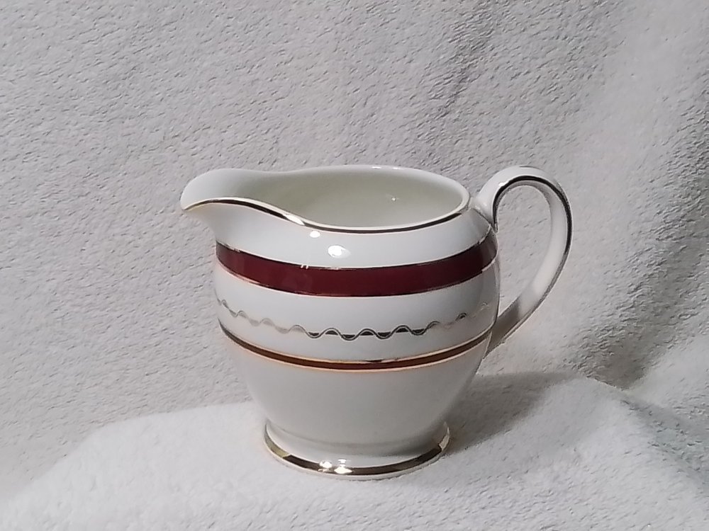 A ceramic creamer with red, black, and gold decorative stripes on a white background.