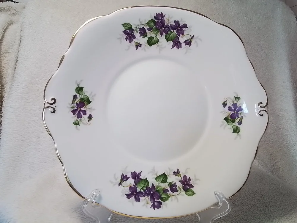 White porcelain plate with purple flower and green leaf pattern and gold trim around the scalloped edge, displayed on a clear stand.