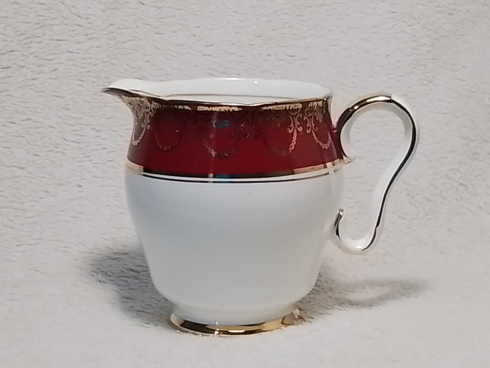 A decorative porcelain creamer with a gold rim, handle, and base, featuring a red and gold ornate pattern near the top.