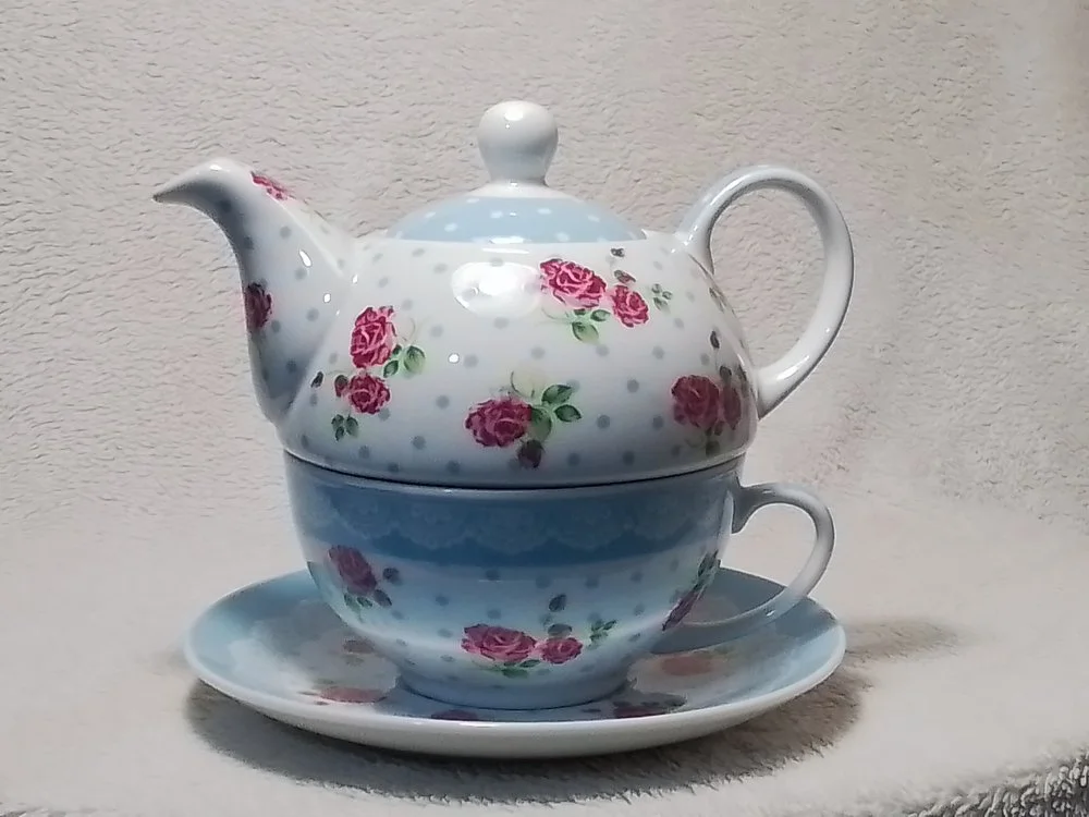 A ceramic tea set with a teapot, a cup, and a saucer, decorated with pink roses and gray polka dots.