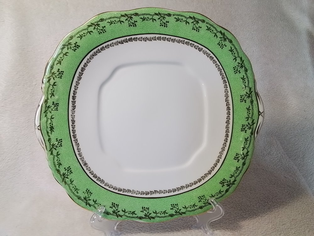 A decorative square white plate with green and black floral design border, displayed on a clear stand.