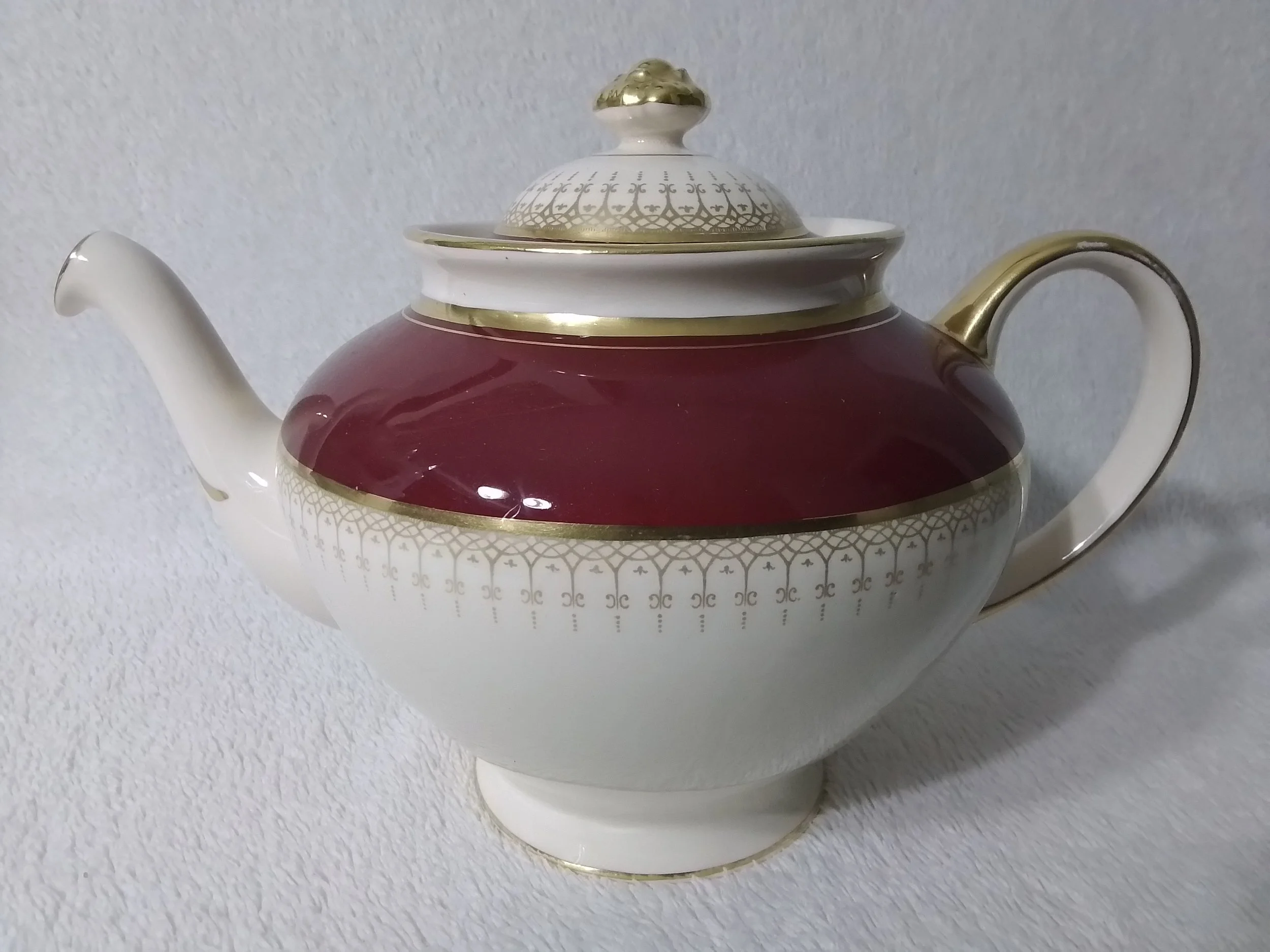 A decorative teapot with white, gold, and burgundy colors, featuring intricate gold patterns.