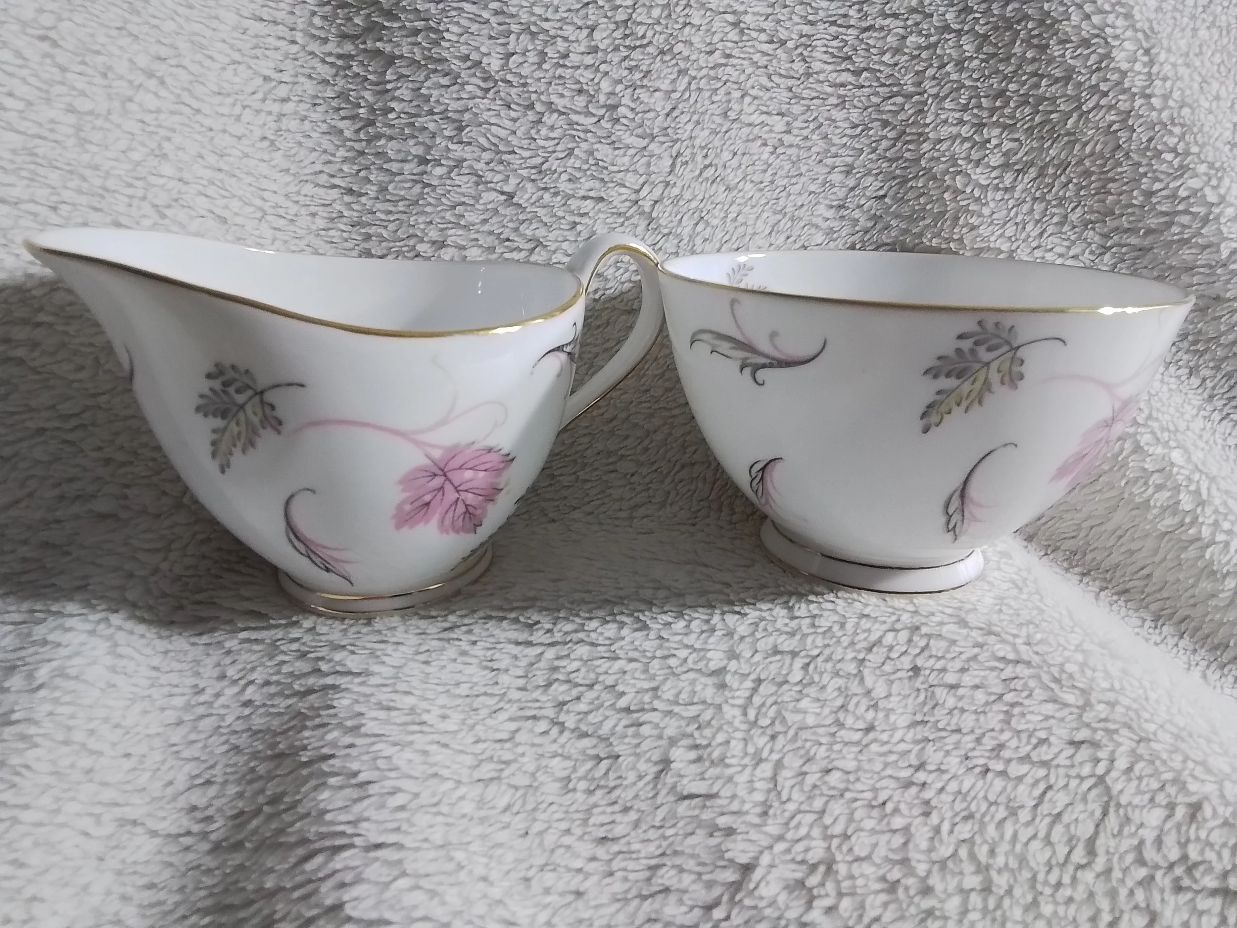 Two white porcelain teacups with floral patterns and gold rims, placed on a soft, textured light gray fabric background.