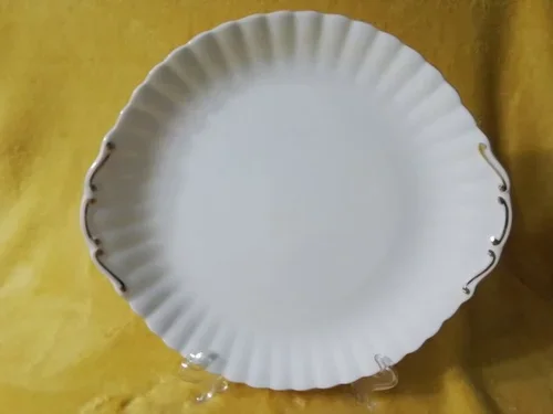 White decorative plate with gold accents on scalloped edges, resting on a small clear stand against a yellow fabric background.