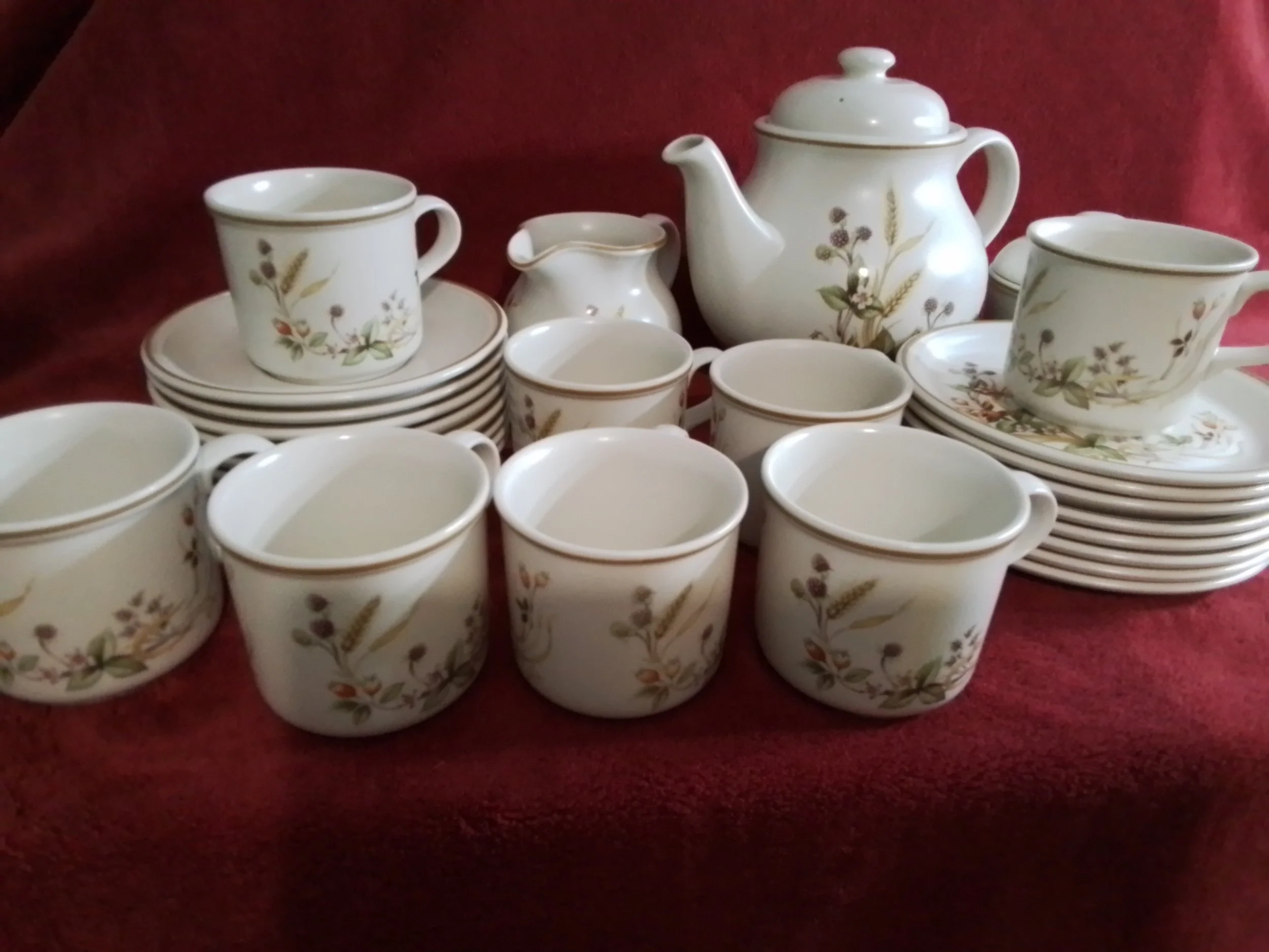 A white ceramic tea set with a floral pattern, including cups, saucers, a teapot, a creamer, and small plates, arranged on a red fabric surface.