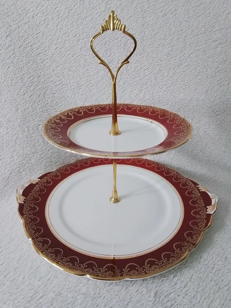 Two-tiered serving tray with a decorative gold handle, red and gold patterned plates, and a white center, placed on a textured light gray surface.