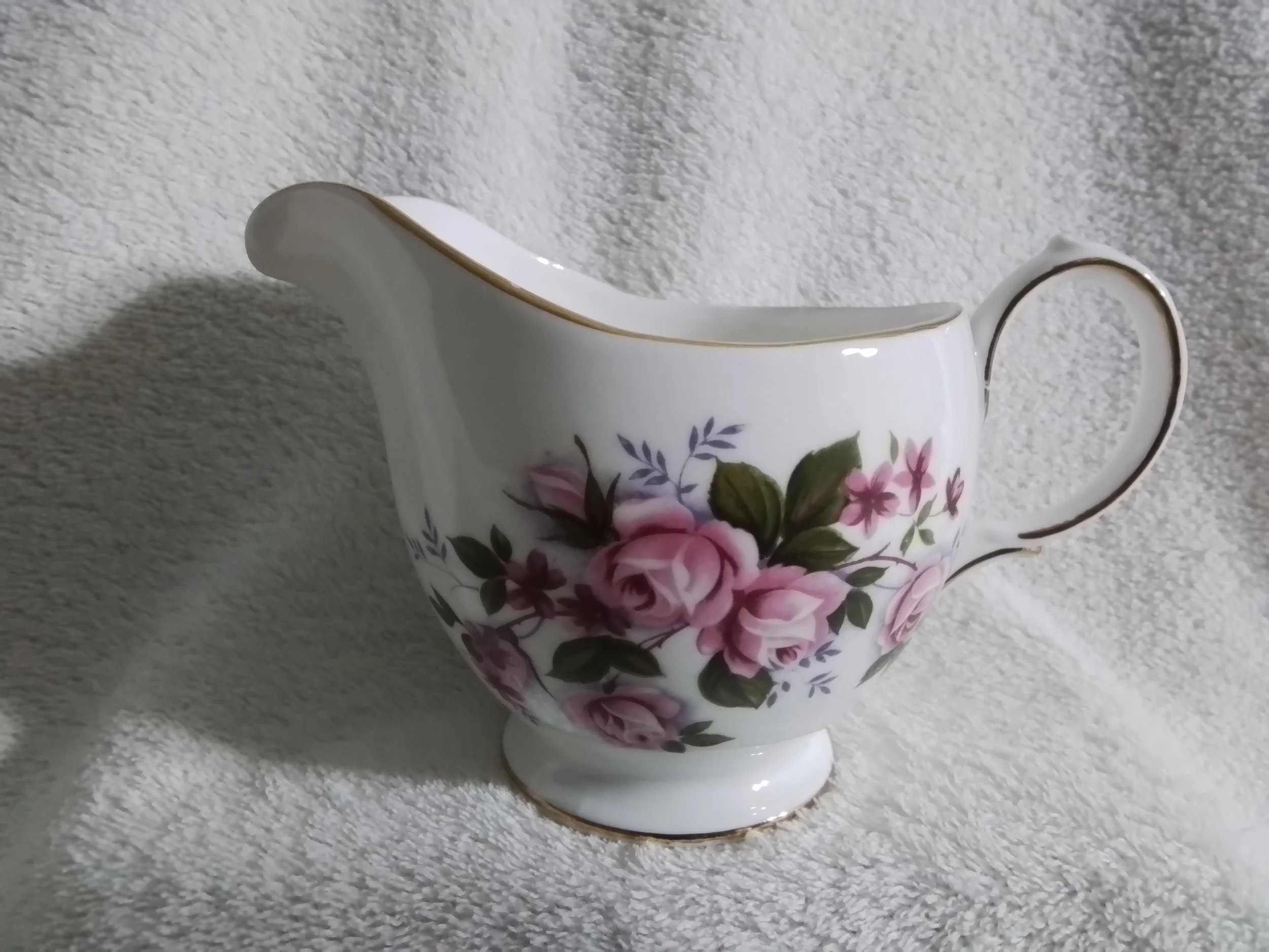 A porcelain creamer with pink and purple floral pattern, gold trim, curved handle, and spout, placed on a textured white surface.