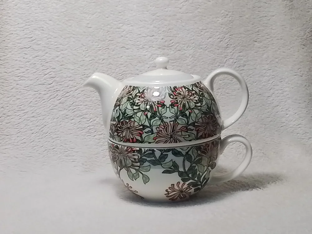 A decorative ceramic teapot with a matching cup, decorated with floral patterns, against a textured white background.