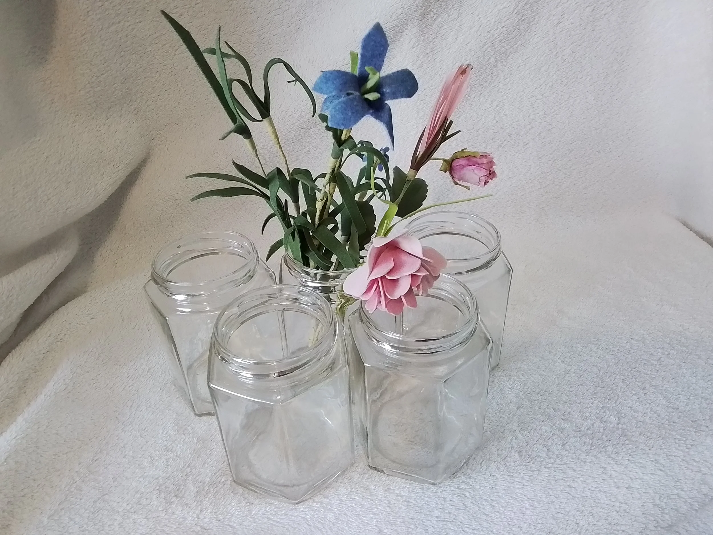 A clear glass vase holding artificial flowers, including pink, blue, and purple blossoms, on a white textured surface.
